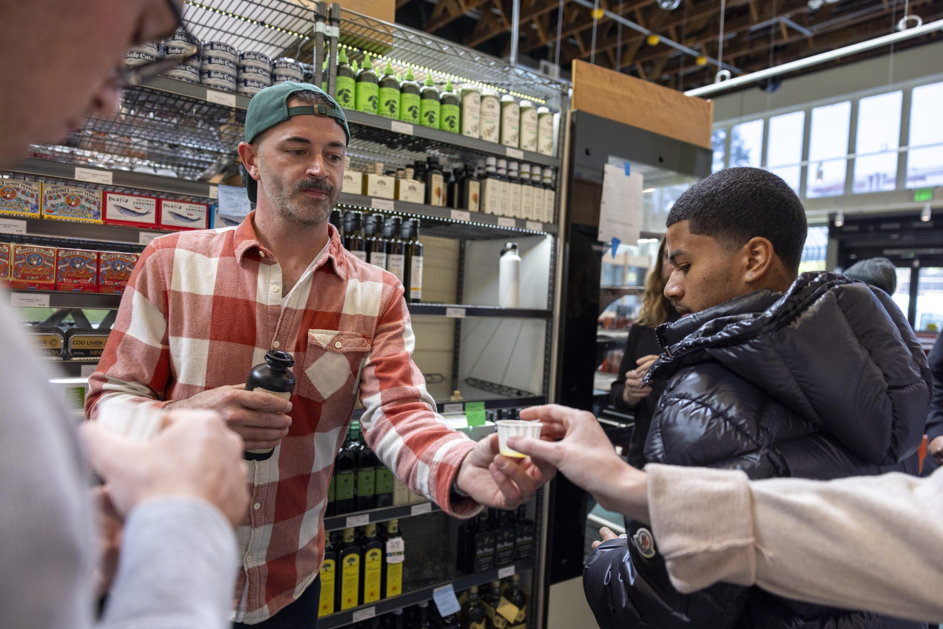 A man in a checkered shirt offers a small cup to another person in a store. Shelves behind them hold various bottles and canned goods.