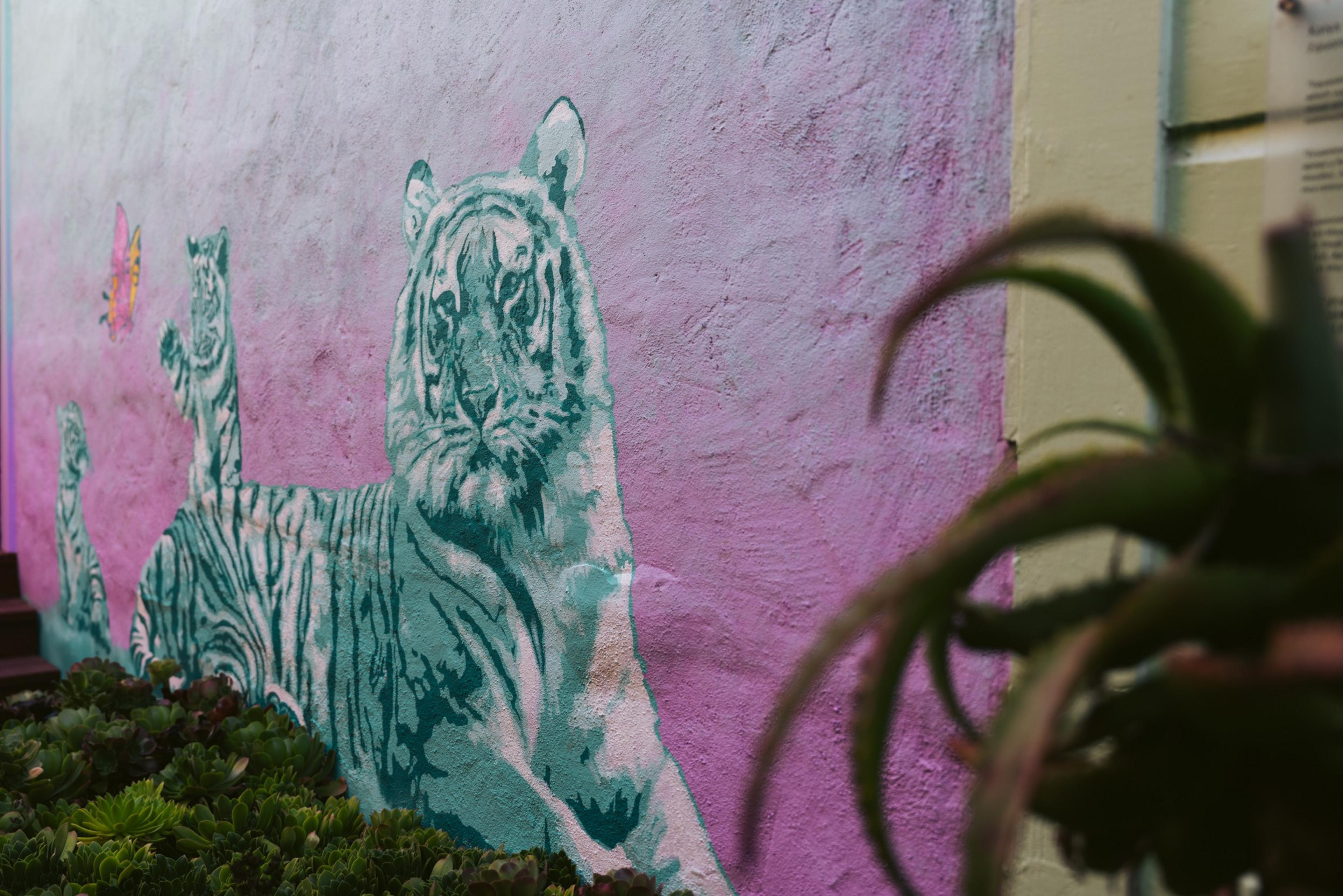 Three greenish-blue tiger cubs are painted on a pink wall, with some green plants growing at the base and a yellow butterfly near one cub.
