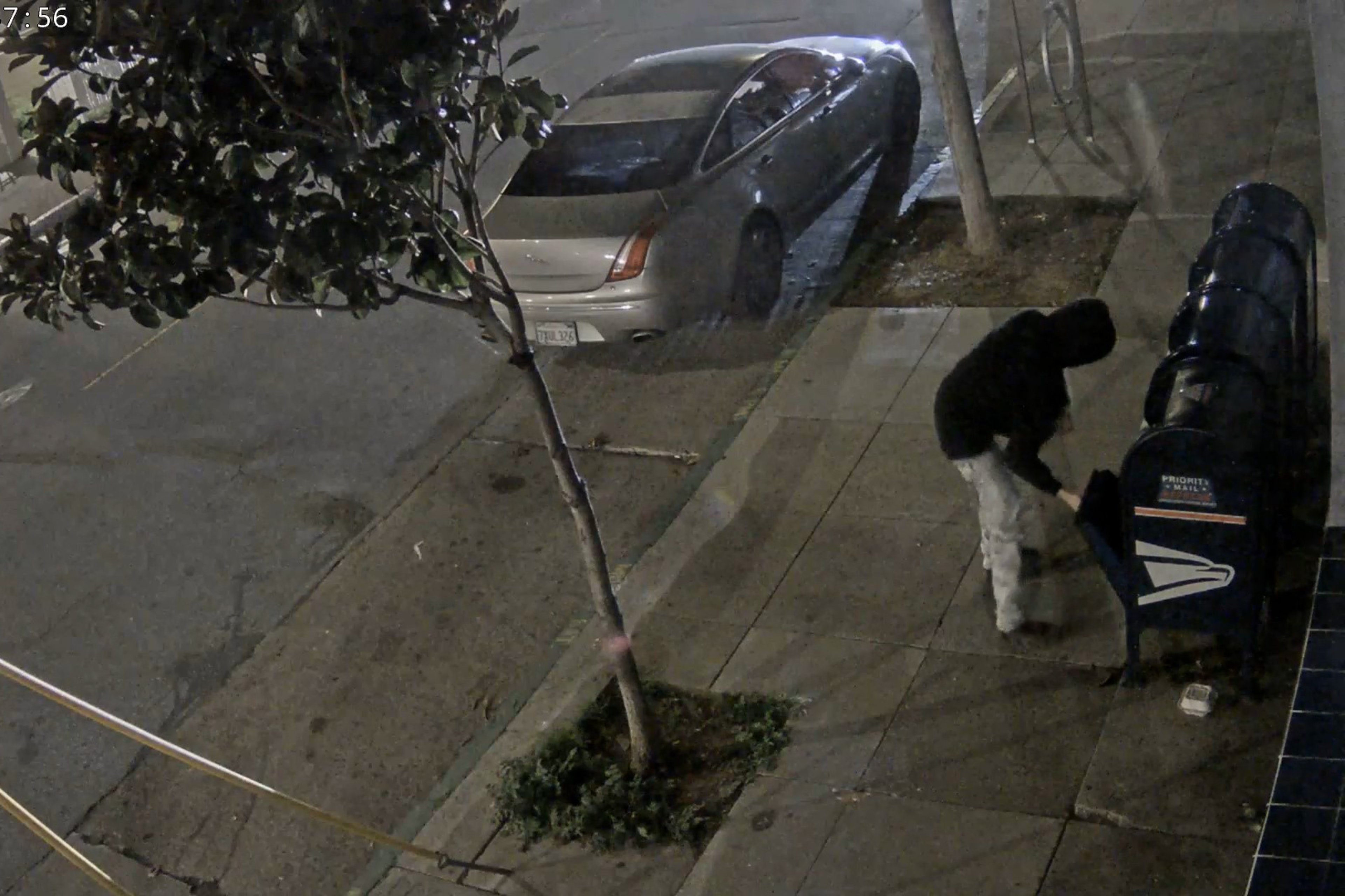 A person wearing a black hoodie is seen on the sidewalk opening blue USPS boxes
