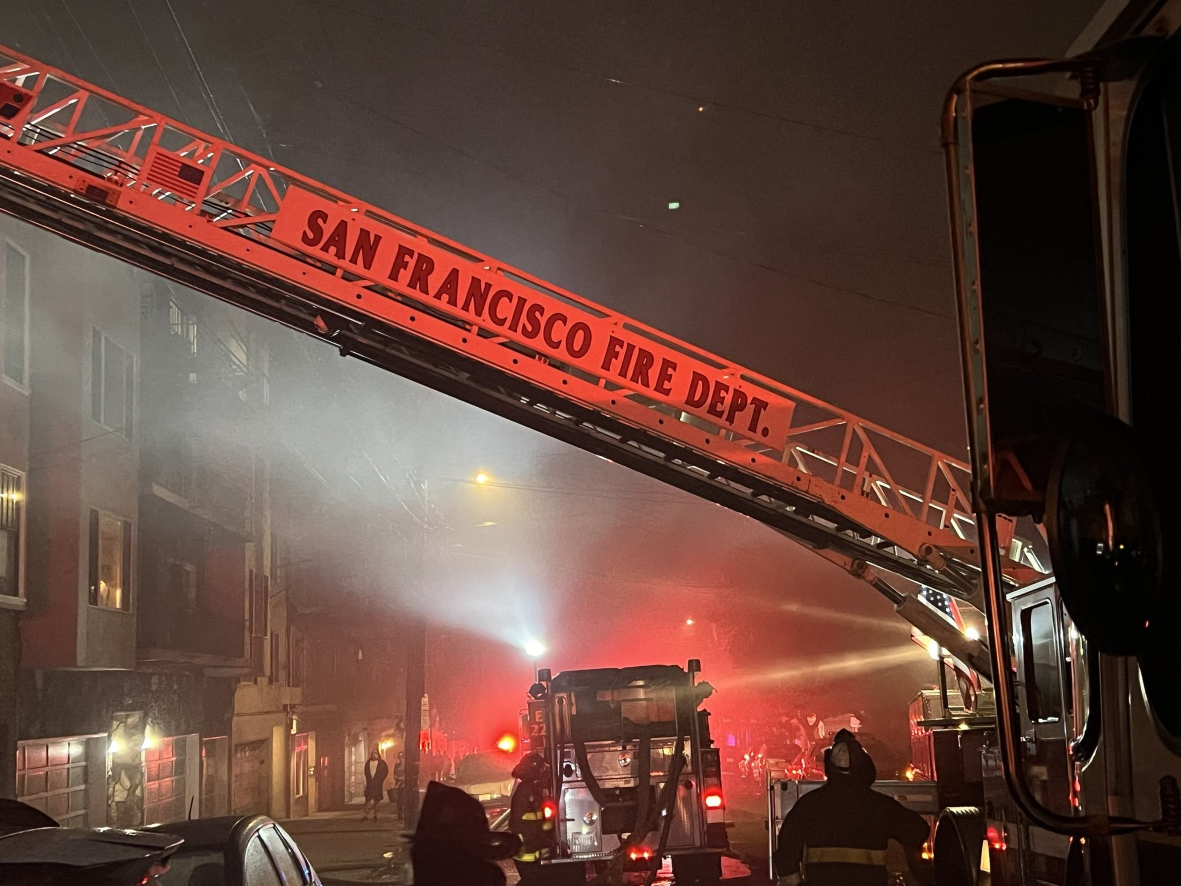 A San Francisco Fire Dept. ladder truck extends over a smoke-filled street with firefighters and emergency lights active at night.