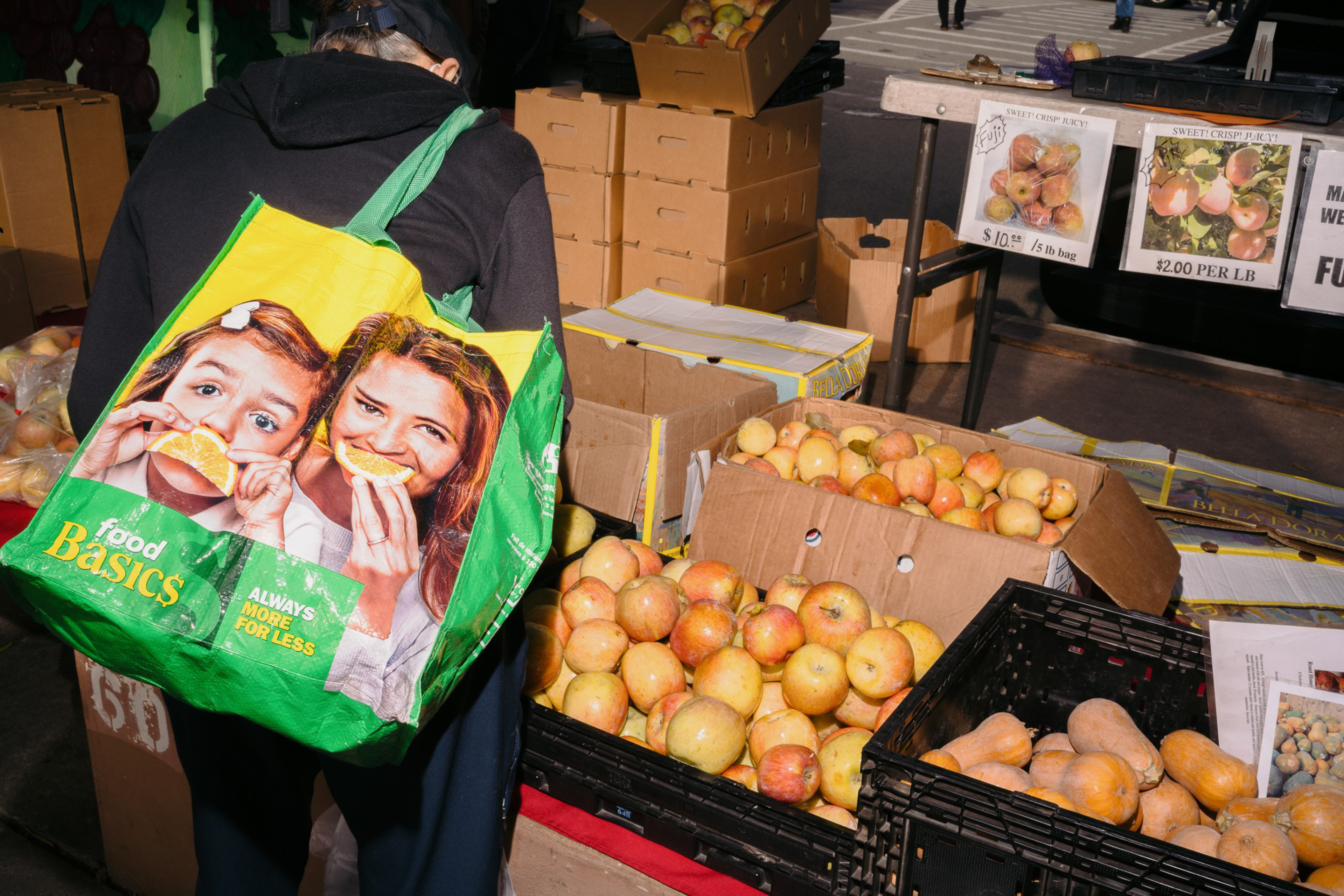 A person with a green tote bag featuring kids eating orange slices stands next to crates of apples and squash at a market stall.