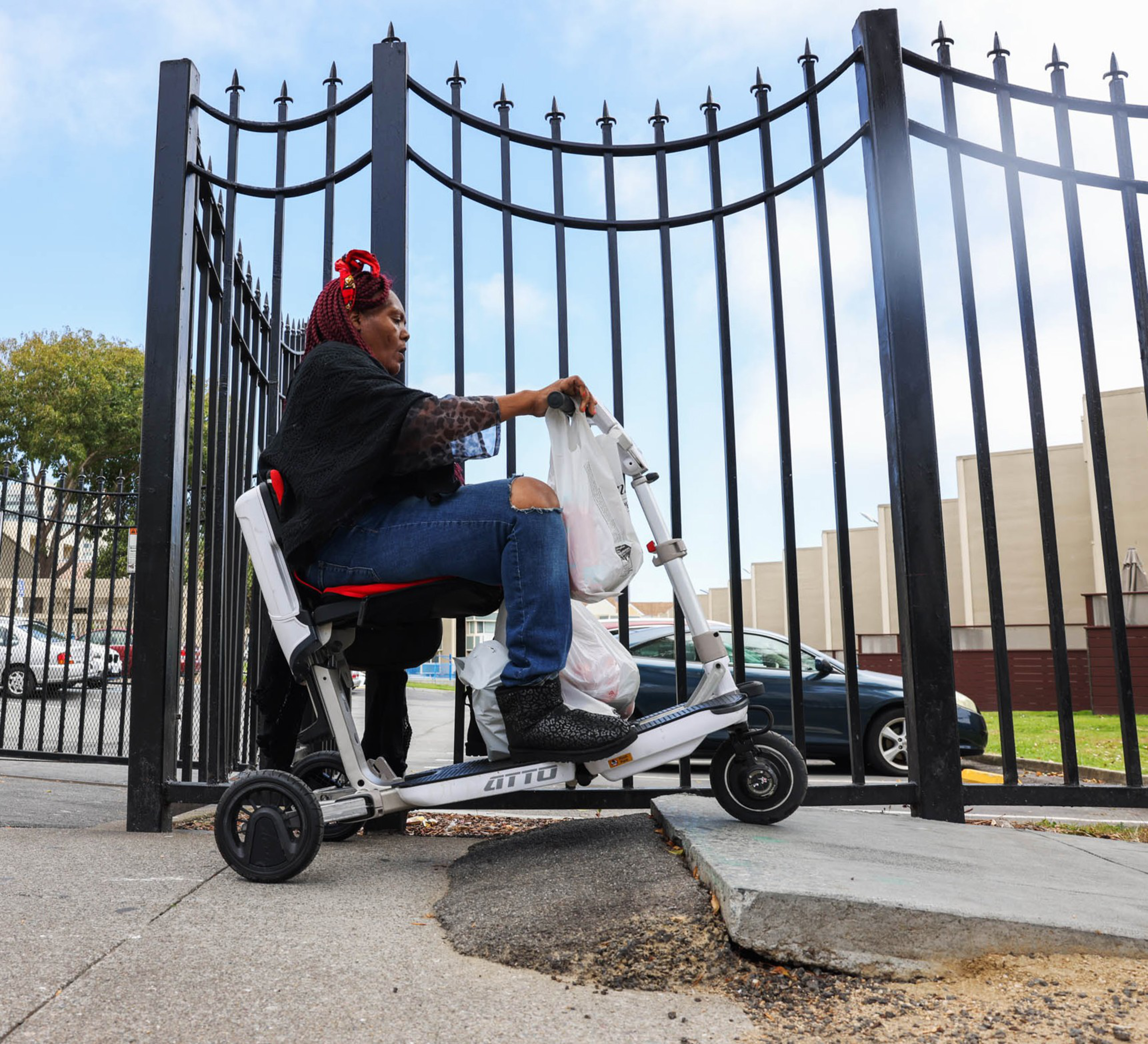 A woman with red hair braids rides a motorized scooter, carrying bags, and navigates up a curb ramp. She's next to a tall black iron fence with a cityscape in the background.