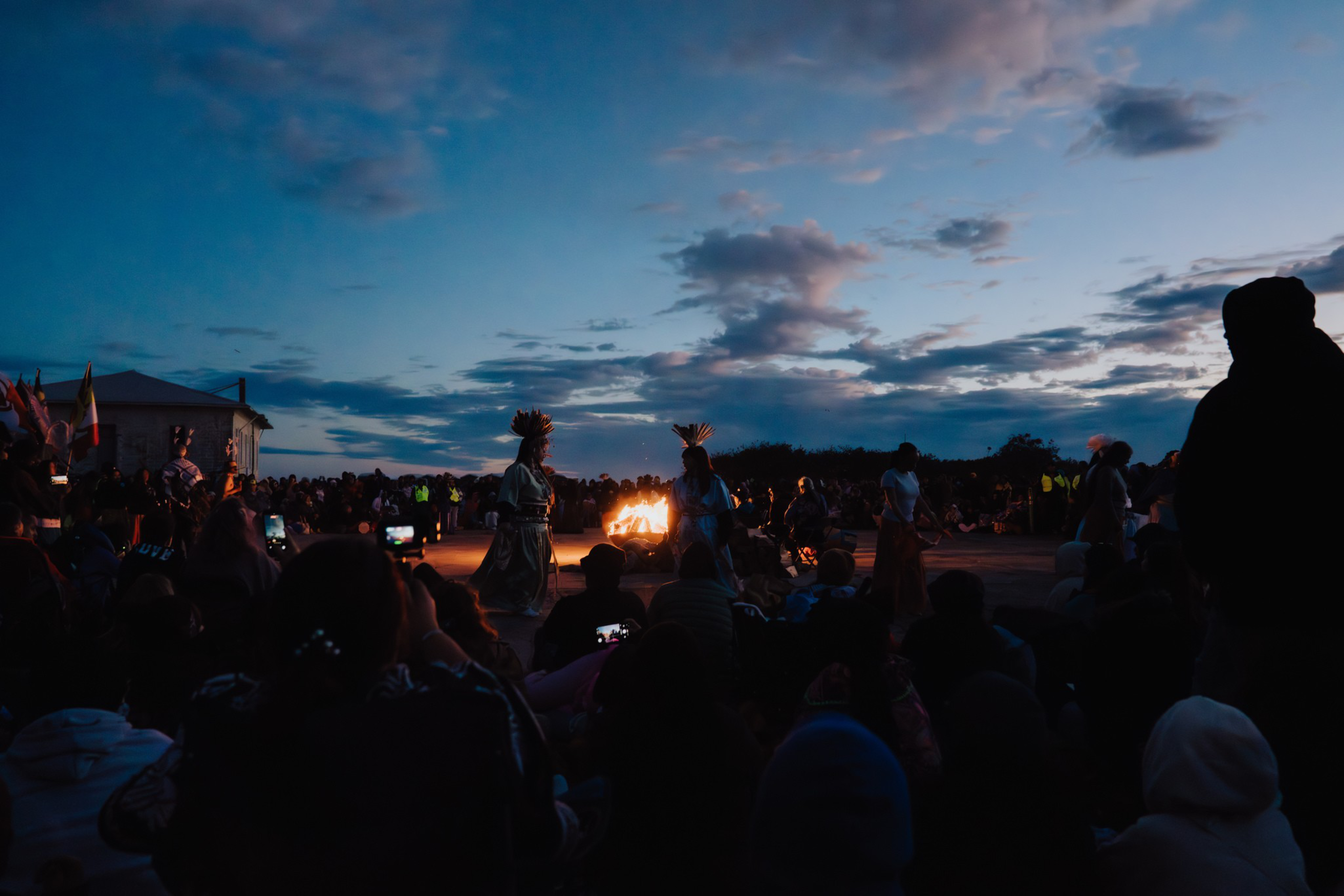 People gather around a fire at dusk, watching performers in traditional attire with headdresses against a darkening sky.