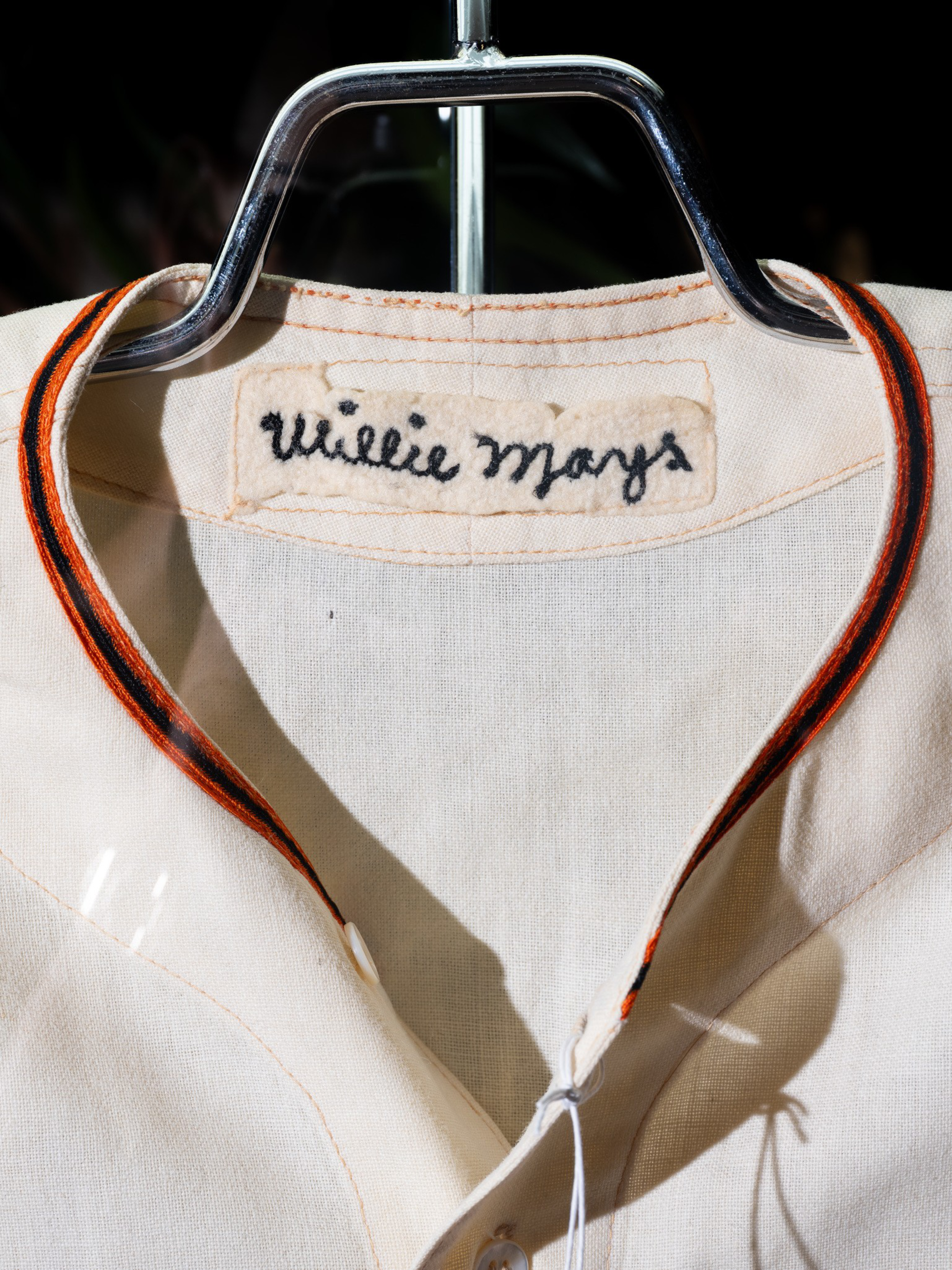 A cream-colored baseball jersey with black and orange trim on the collar, hanging on a metal hanger with “Willie Mays” stitched on a fabric label inside.