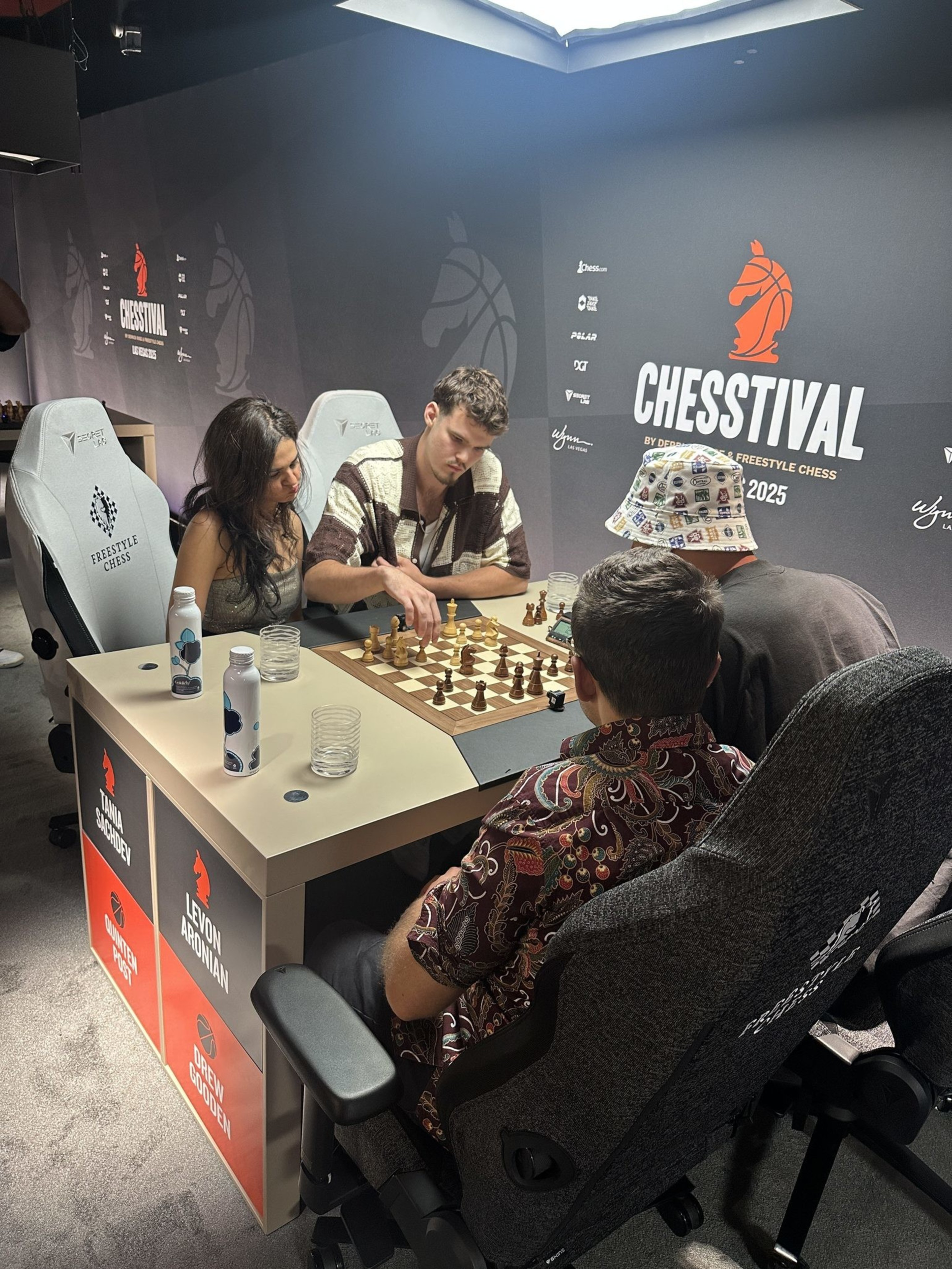 Three people are focused on a chess game at a tournament called Chesstival, sitting in comfy chairs with drinks nearby, under bright lighting.