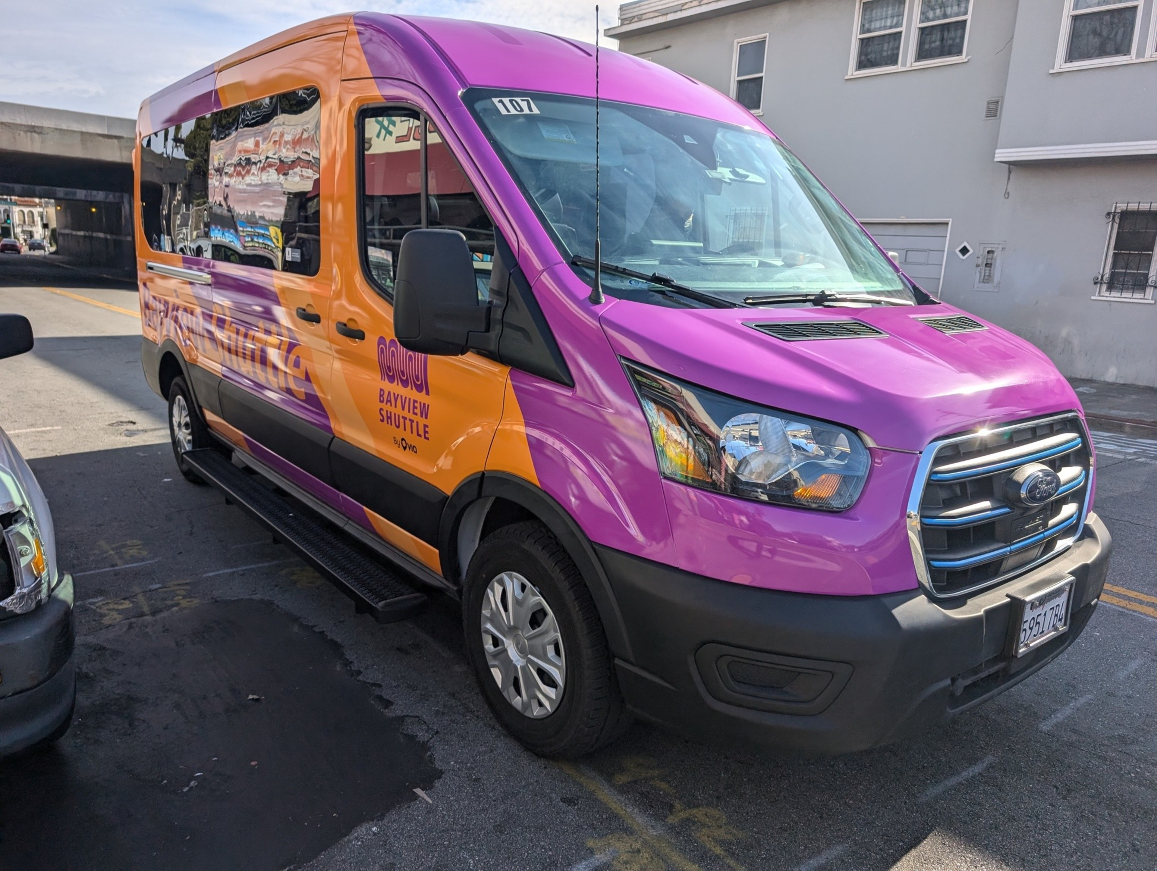 The image shows a vibrant shuttle van with a purple and orange color scheme, parked on a street. It displays &quot;Bayview Shuttle&quot; and &quot;aqui&quot; on its side.