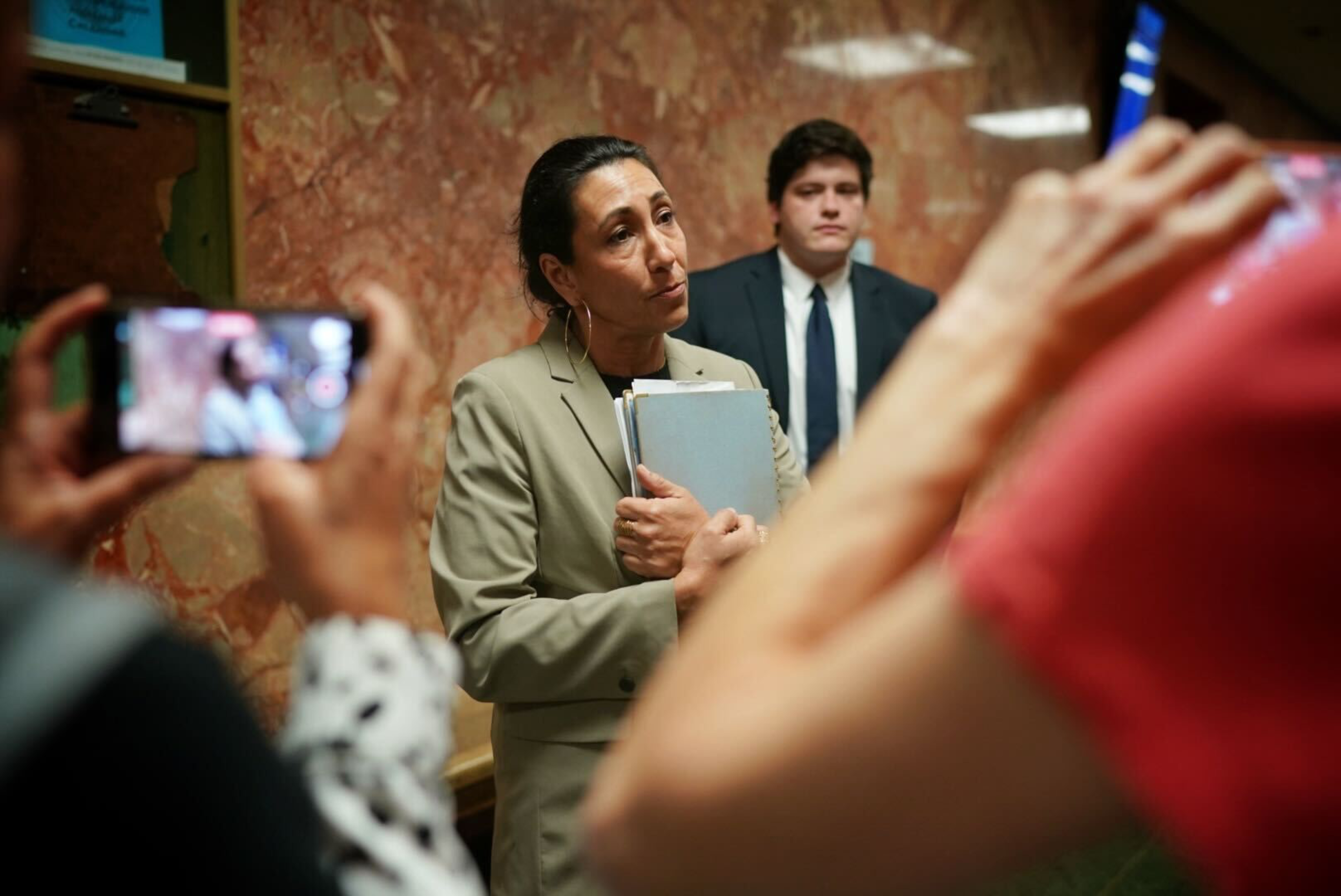 A woman holding documents speaks to other people, one that is recording her on a mobile phone.