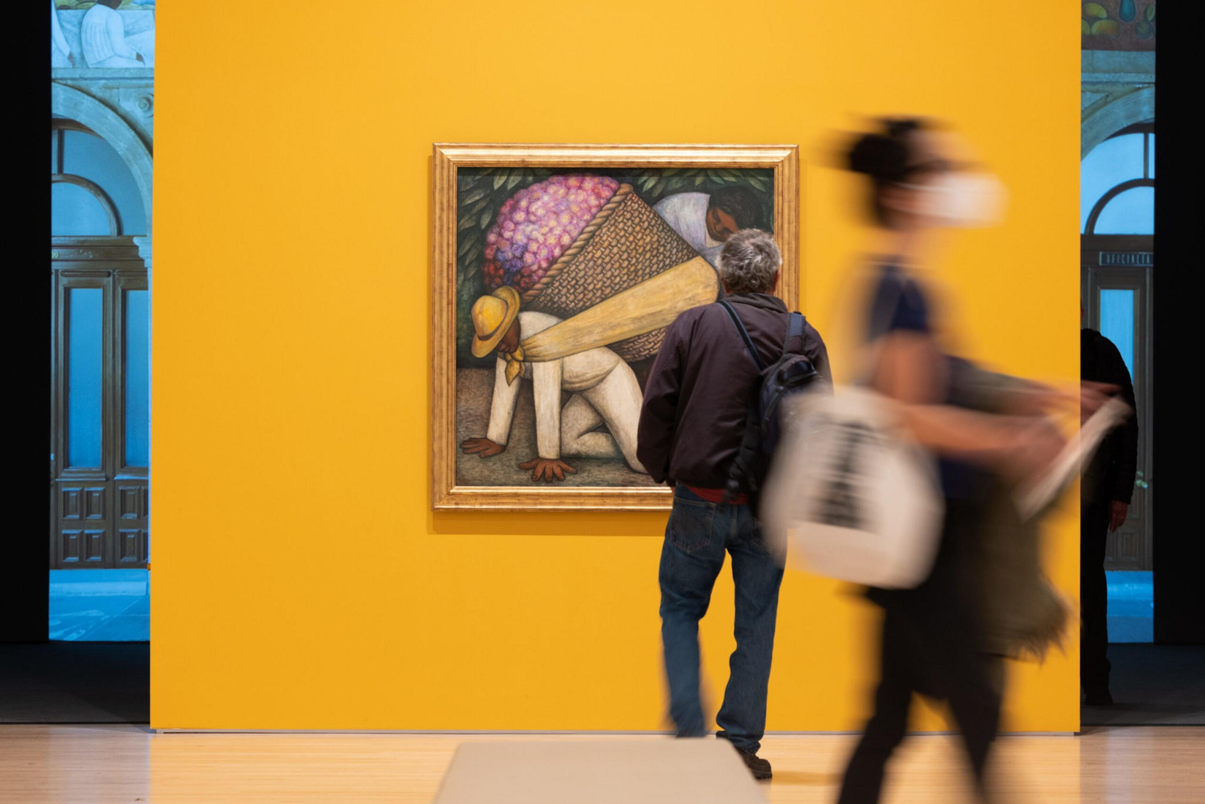 A person stands closely examining a vibrant painting on a yellow wall, depicting a figure carrying a large basket of purple flowers. Another person walks by, blurred.