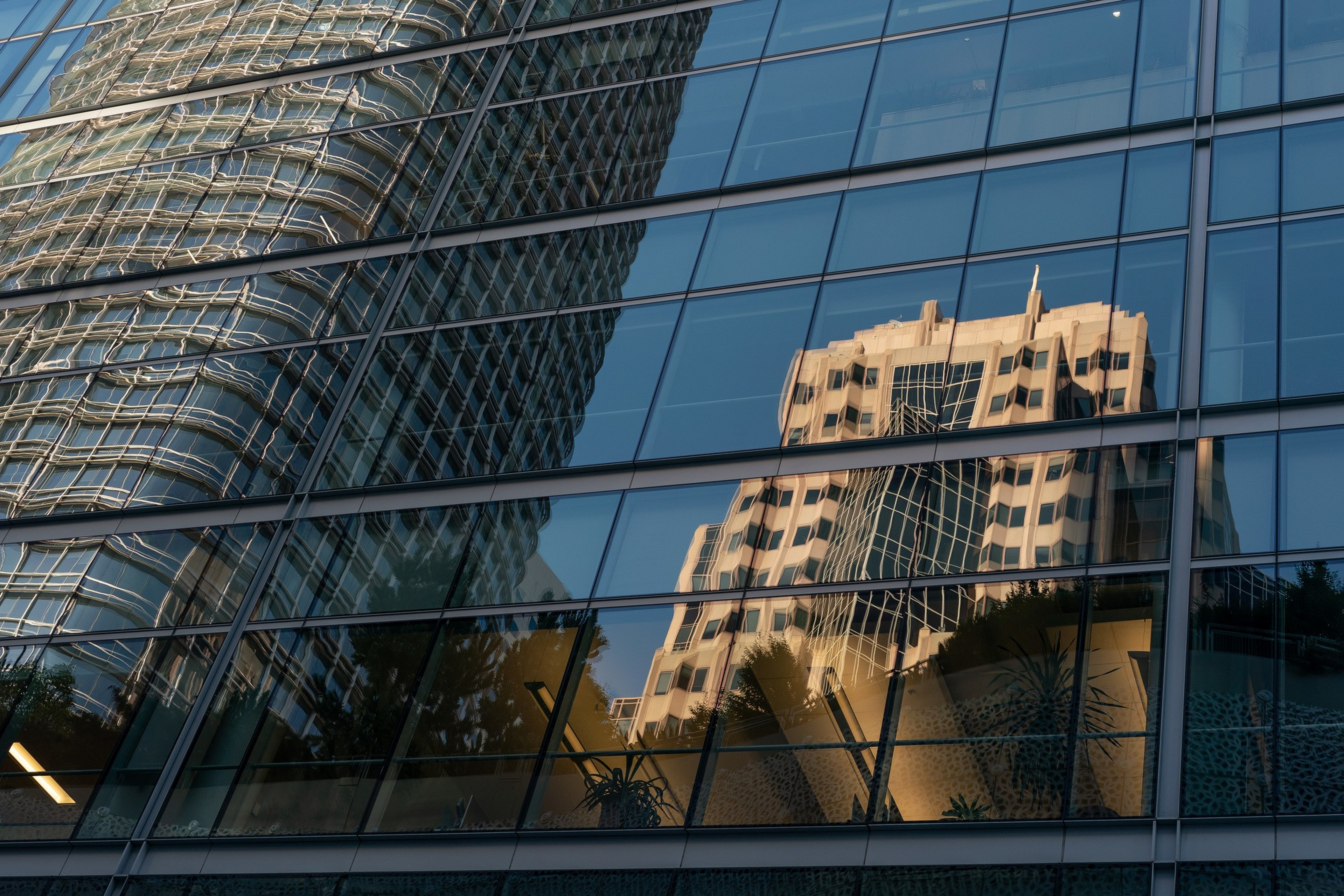 Two tall buildings are reflected in the glass windows of another building, showing distorted and mirrored architectural details.