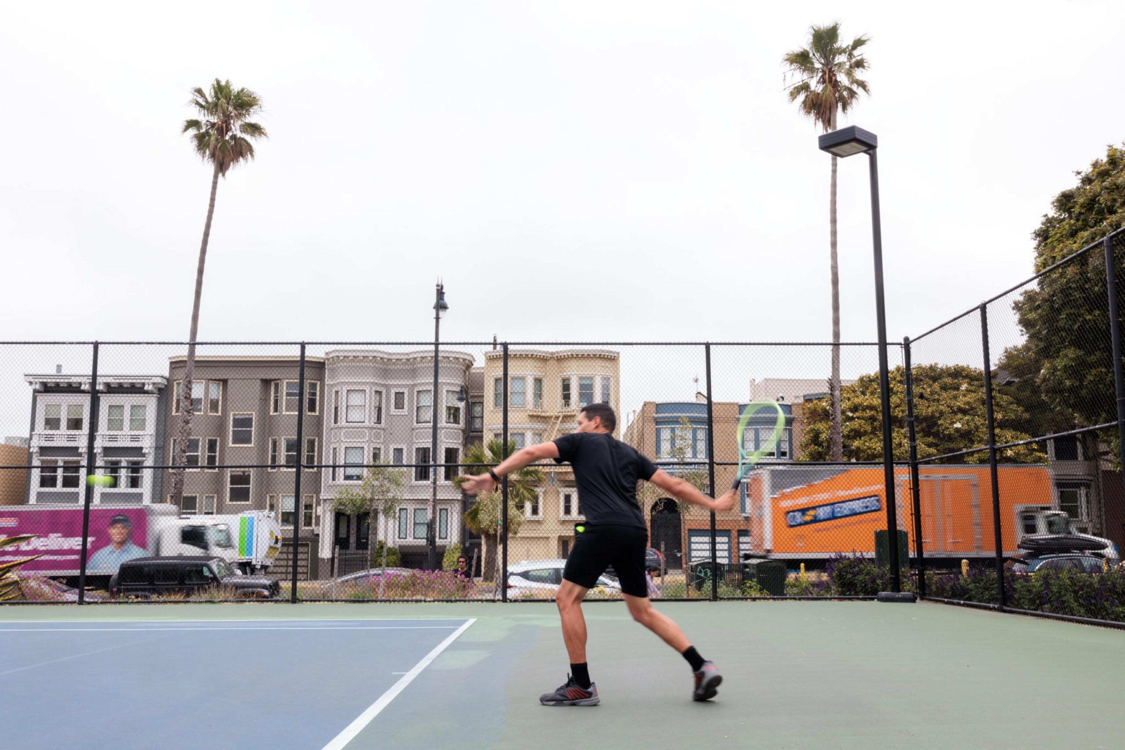 A man dressed in black prepares to hit a tennis ball on an outdoor court, with palm trees, colorful buildings, and passing trucks in the background.