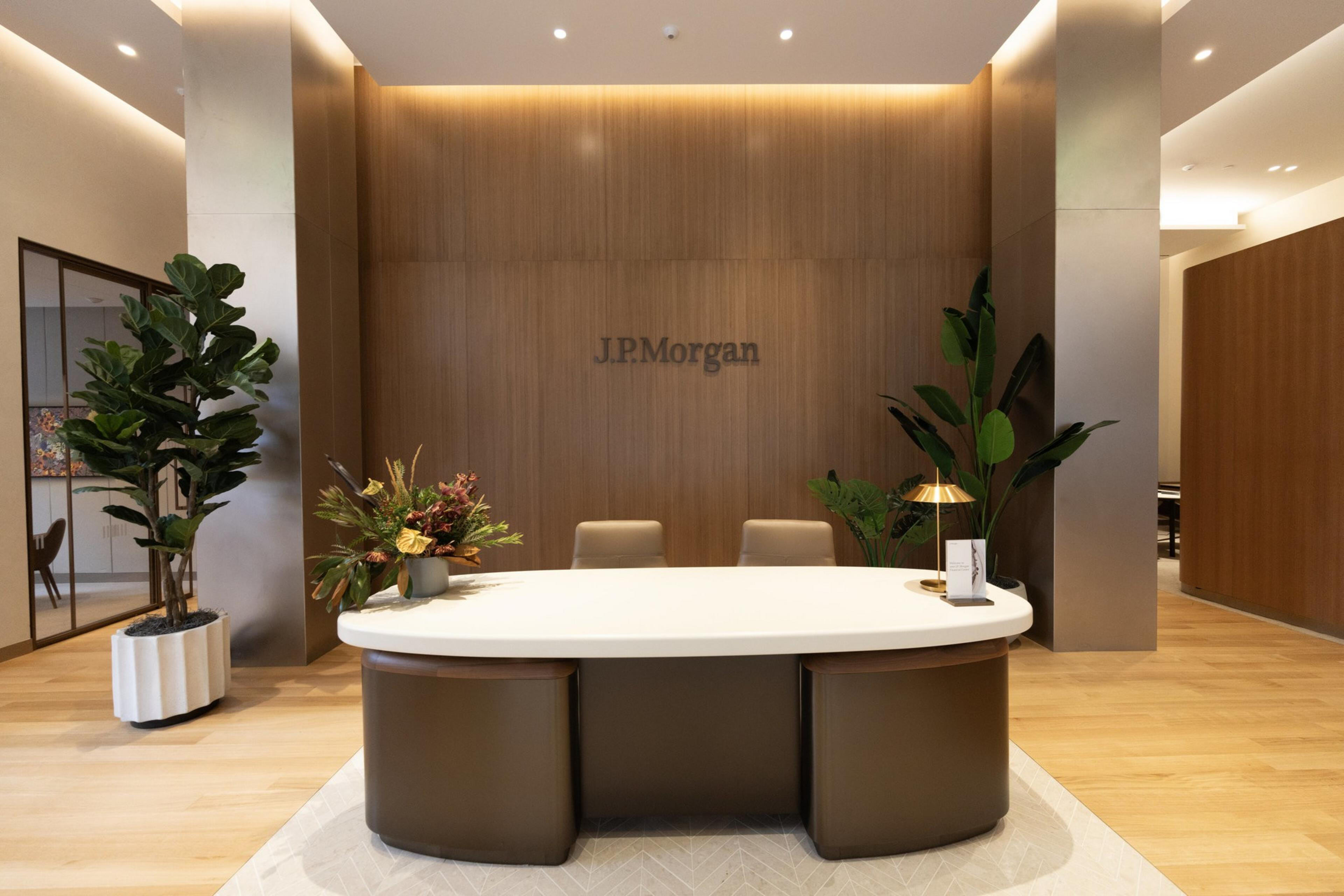 The image shows a modern office reception area with a wooden wall displaying the J.P. Morgan logo, a sleek white desk, chairs, plants, and soft lighting.