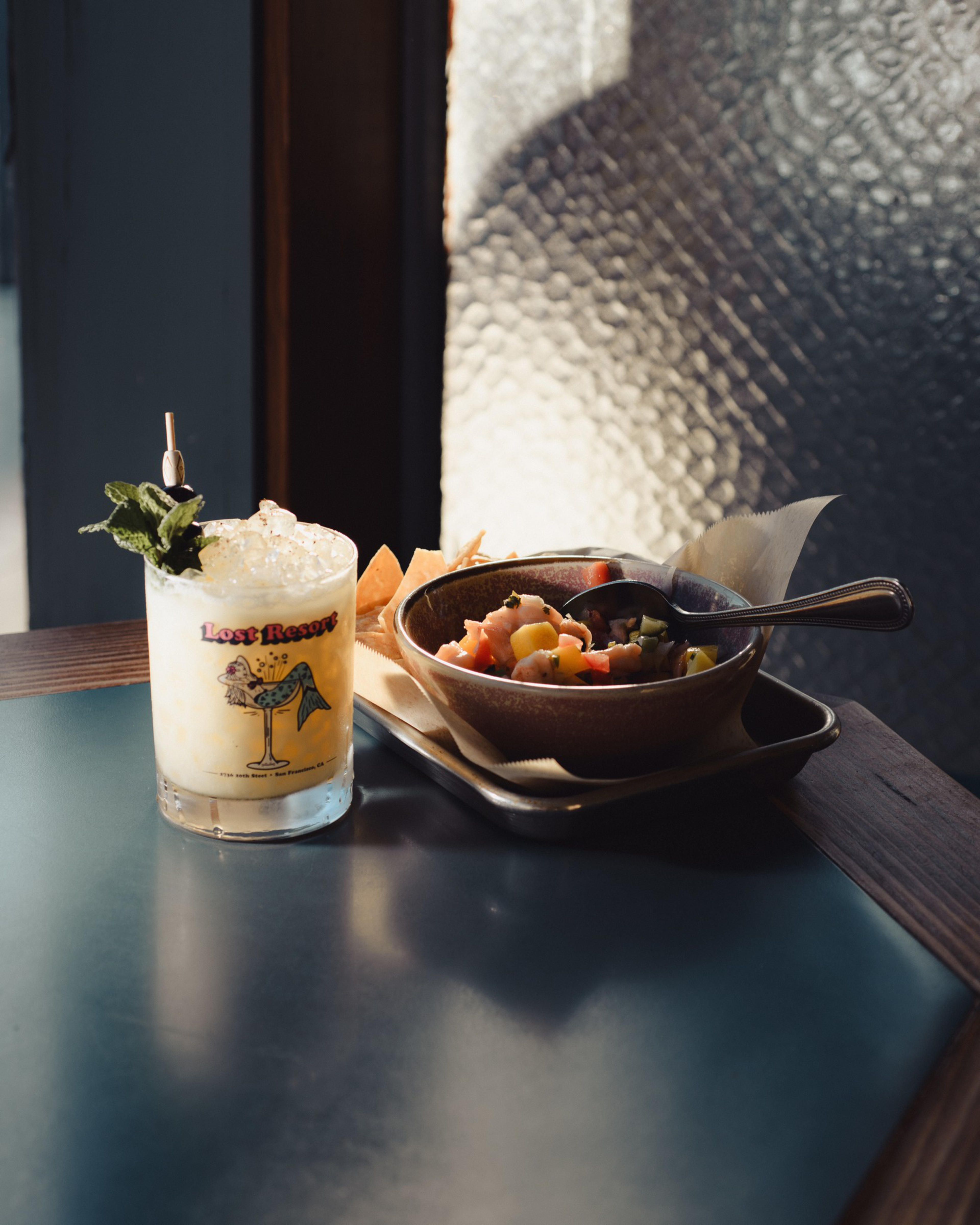 A creamy cocktail garnished with mint sits next to a bowl of mixed seafood ceviche and tortilla chips on a tray, all on a dark tabletop.