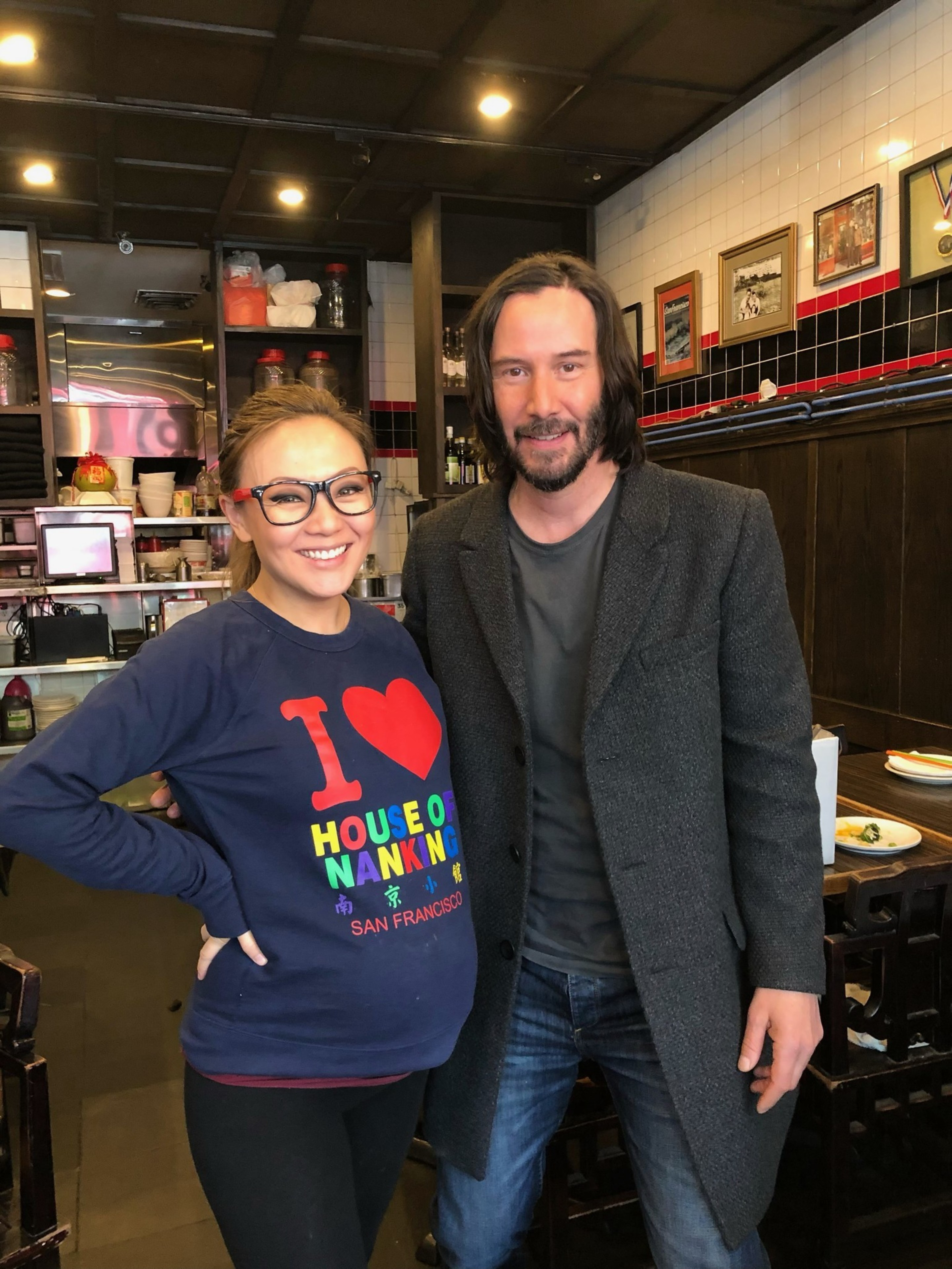 A smiling woman wearing glasses and a colorful “I ♥ House of Nanking” sweatshirt stands next to a man with shoulder-length hair and a dark coat in a restaurant.
