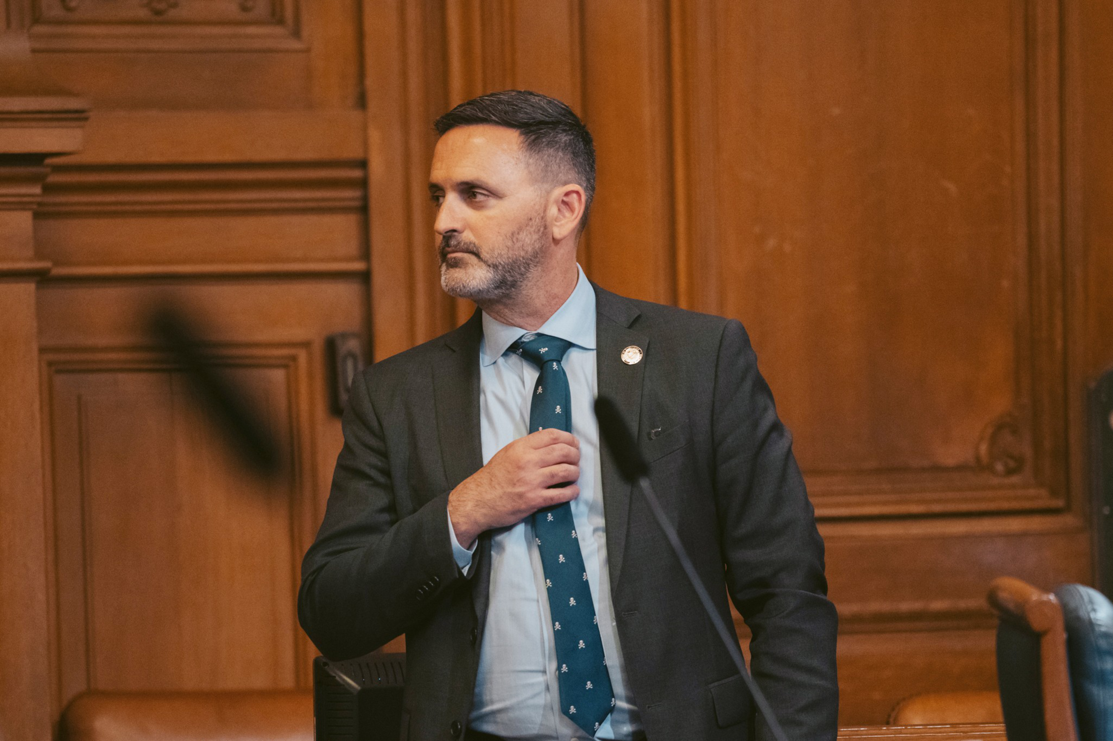 A man in a dark suit and teal tie adjusts his tie while looking to his left in a wood-paneled room with microphones in front of him.