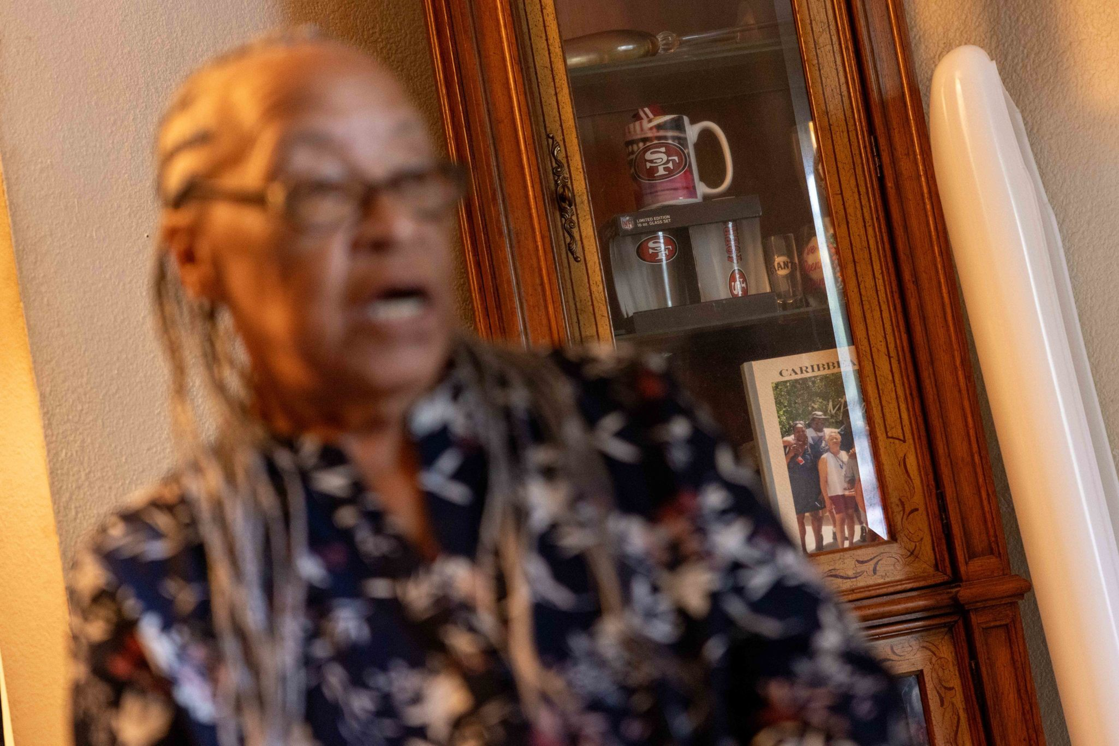 An older person with braided hair and glasses wears a floral shirt, standing near a wooden cabinet holding San Francisco 49ers mugs and a framed photo.