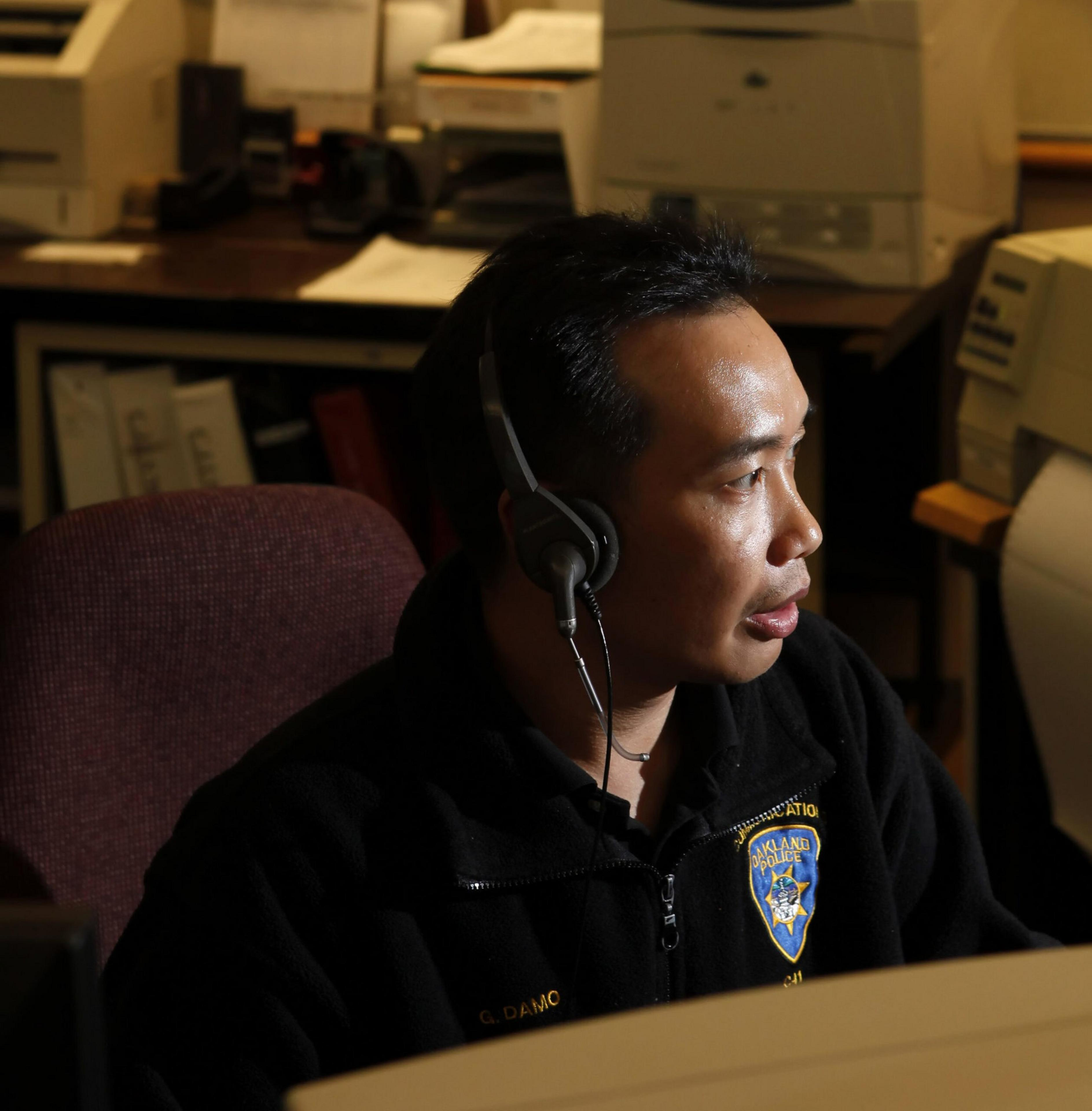 A man wearing a black jacket with an Oakland Police badge and a headset is seated at a desk in an office with printers and papers.