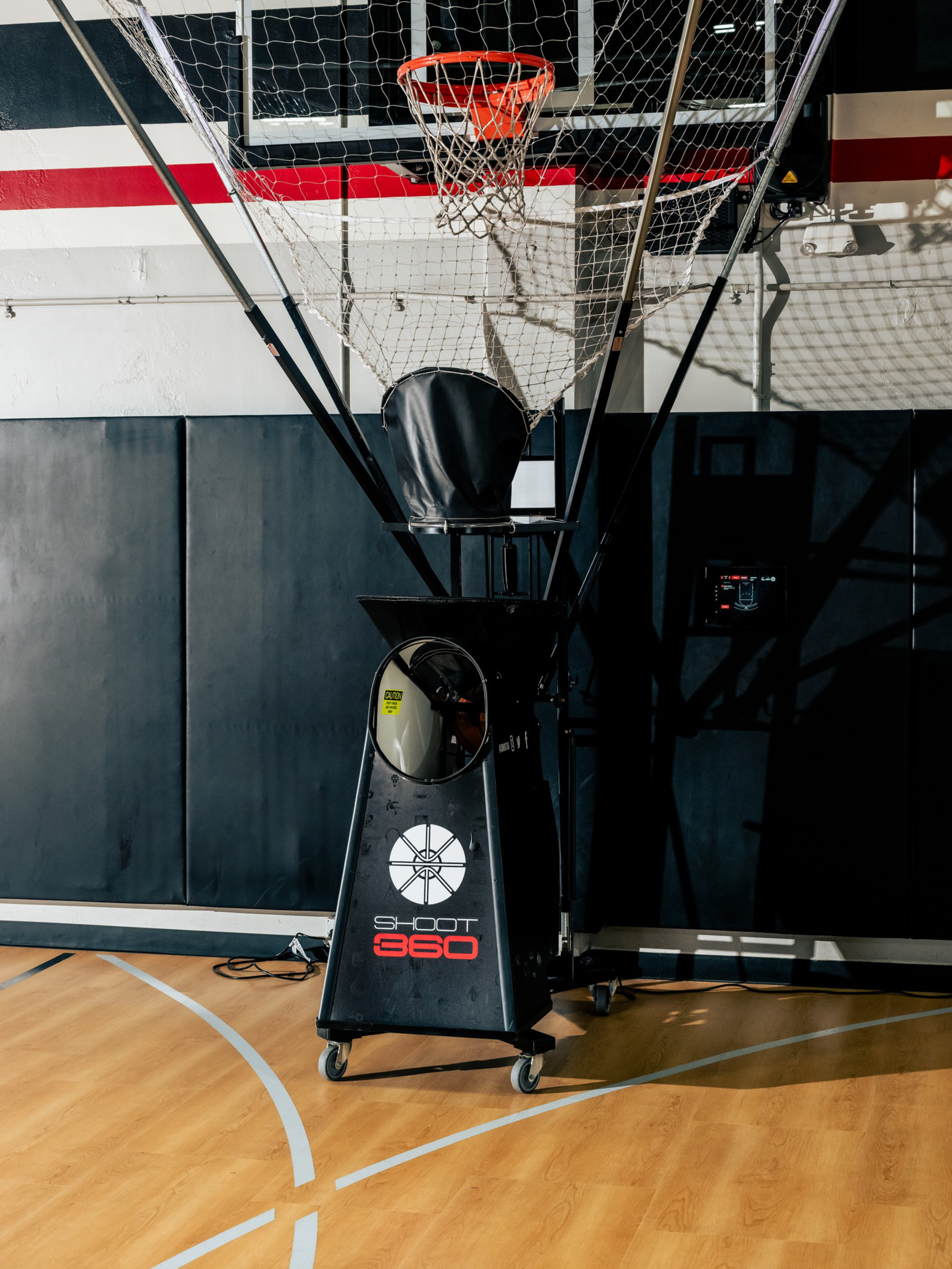 A Shoot 360 basketball shooting machine with a hoop, netting, and a black base on wheels stands on a gym basketball court.