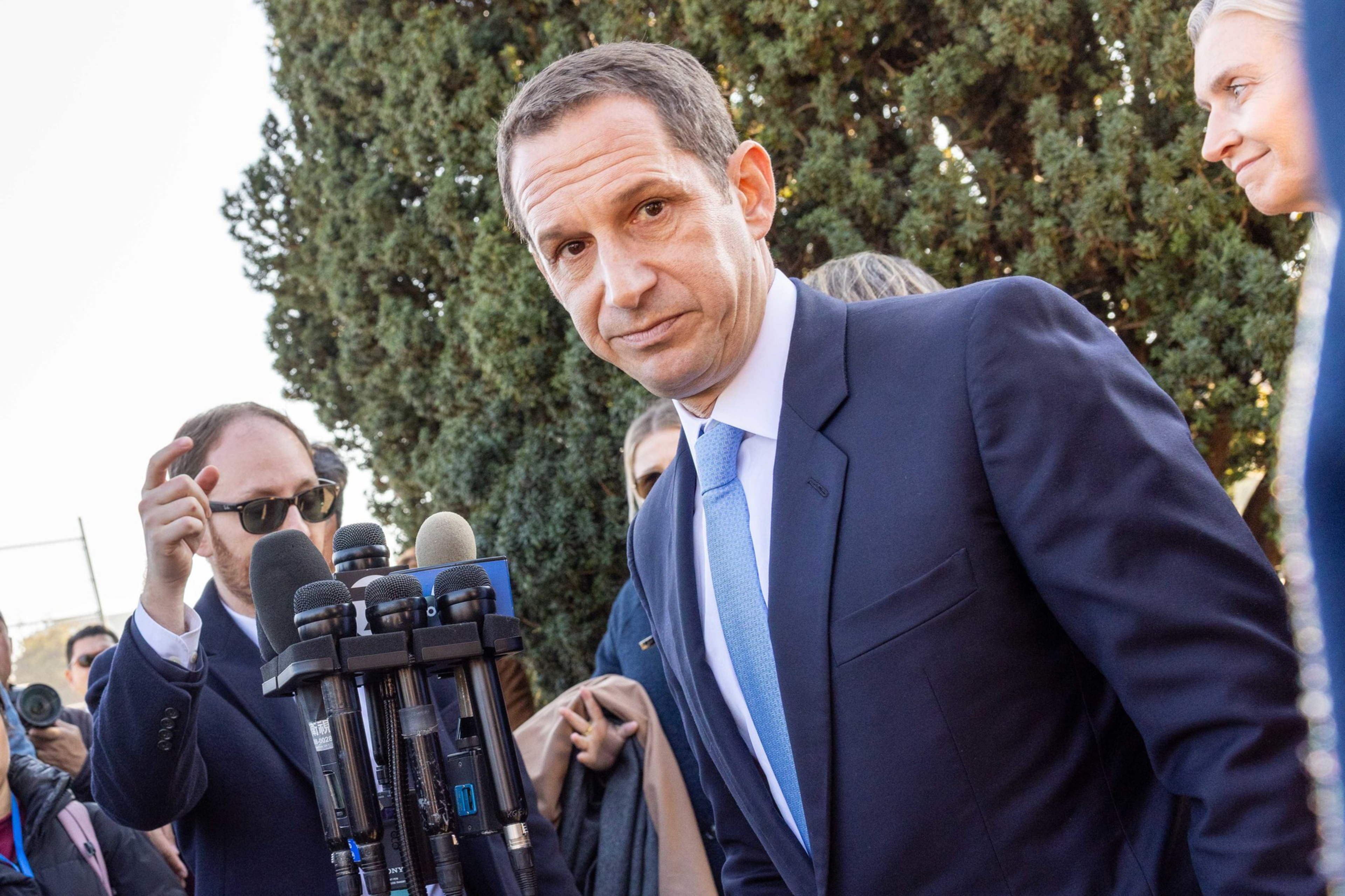 A man in a blue suit and light blue tie leans toward several microphones, while another man wearing sunglasses and a group of people stand behind him.