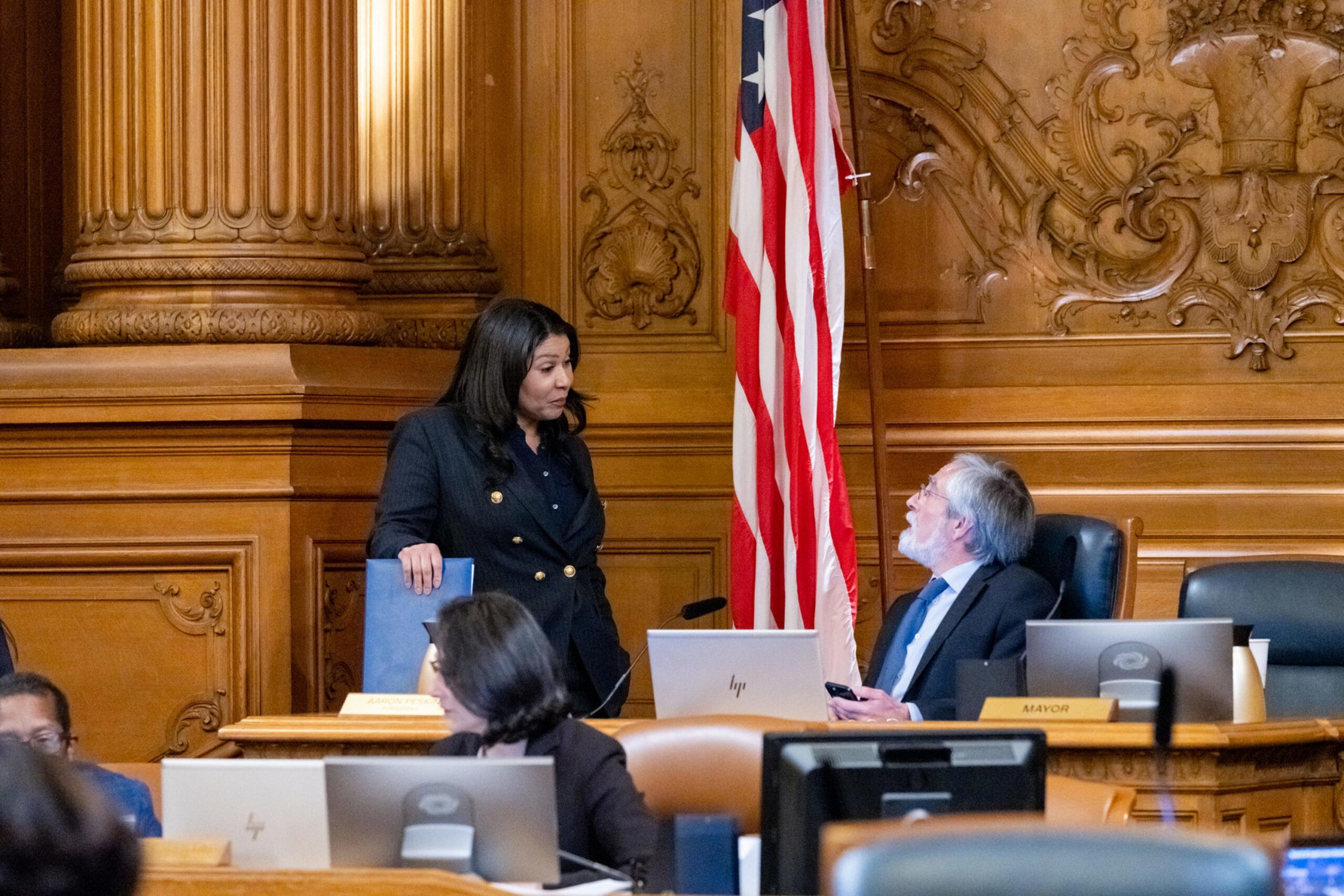The image depicts two people in a formal setting with wood paneled walls and an American flag. The woman is standing, holding a folder, and talking to a seated man.