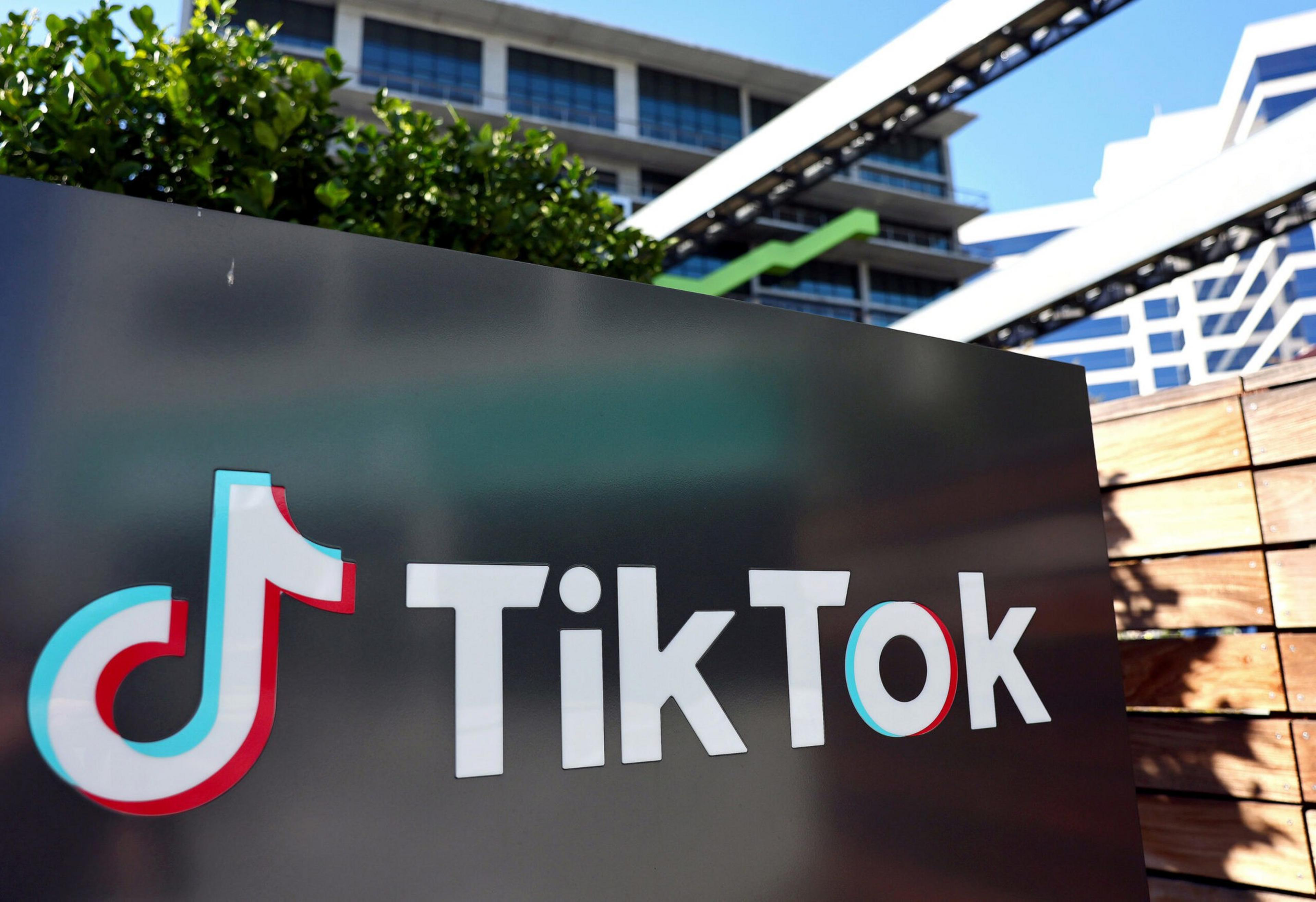 A black sign displays the TikTok logo and name in white letters with a blue and red outline, set outdoors near modern buildings and greenery.