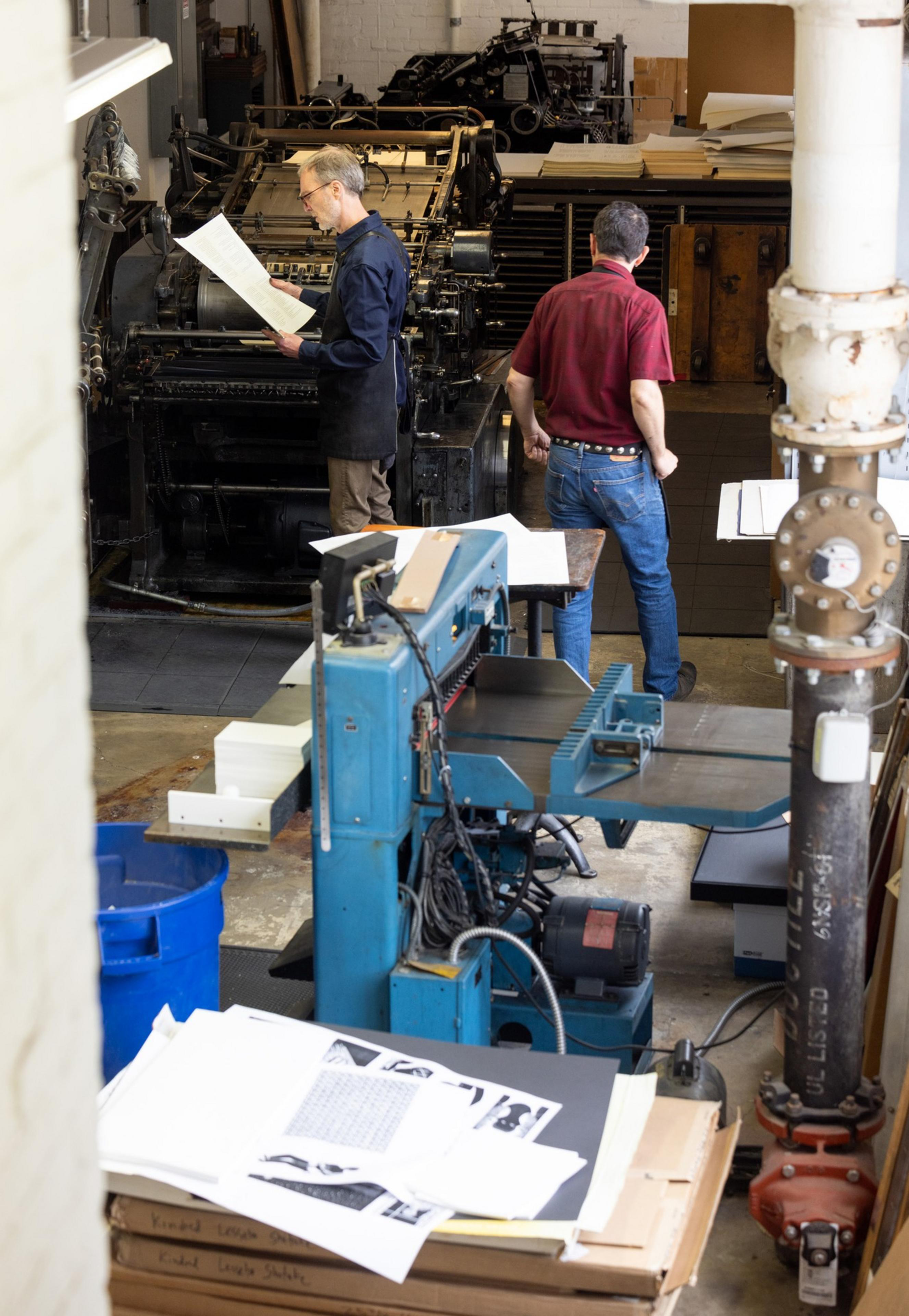 Two men work in a busy print shop filled with large machines and papers, checking the quality of freshly printed sheets.
