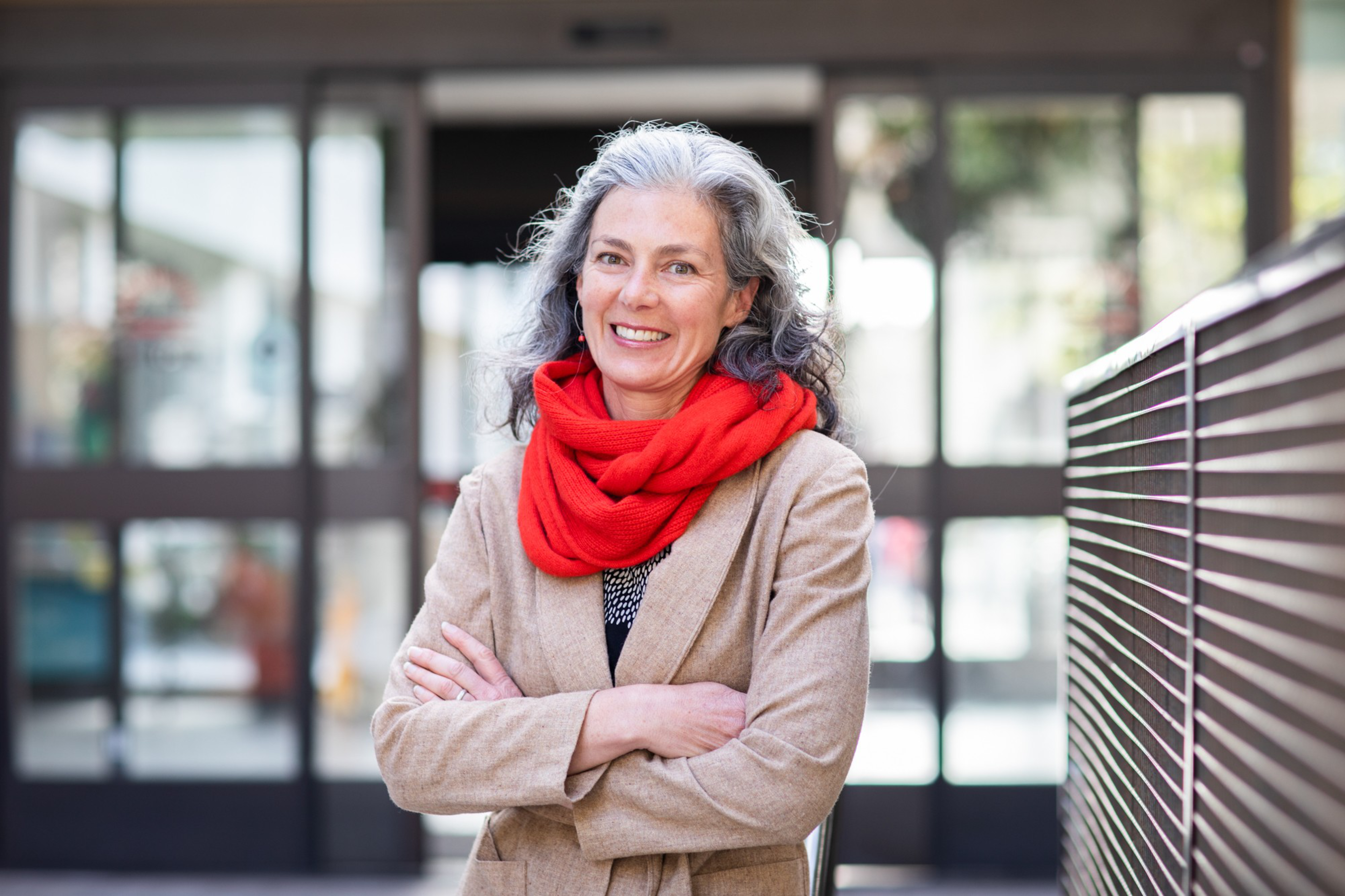 A smiling middle-aged woman with curly gray hair wears a red scarf and beige coat, standing with arms crossed in front of blurred glass doors.