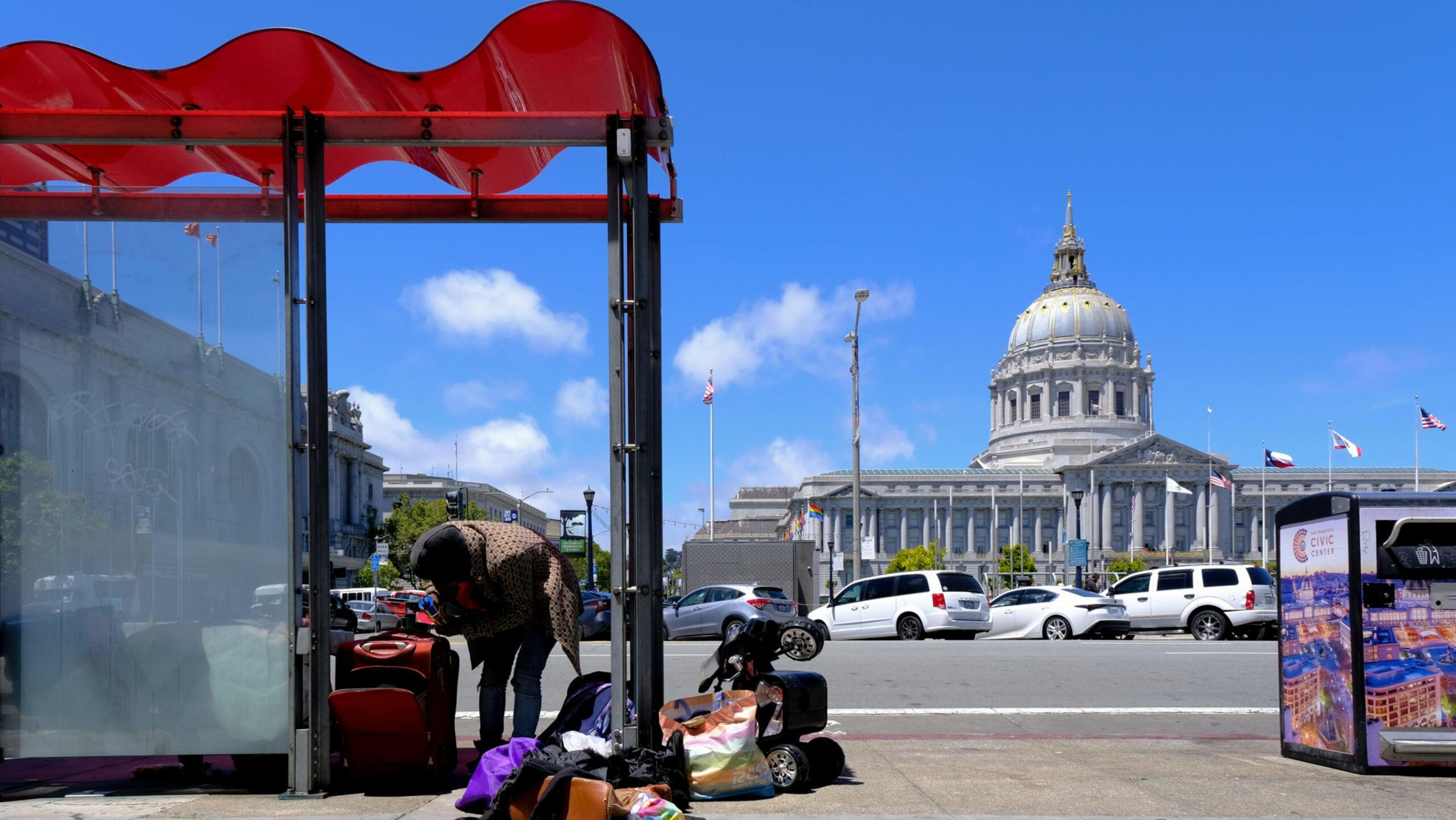 A person bends over next to a bus stop with luggage and bags scattered around under a red canopy, with a domed government building in the background.