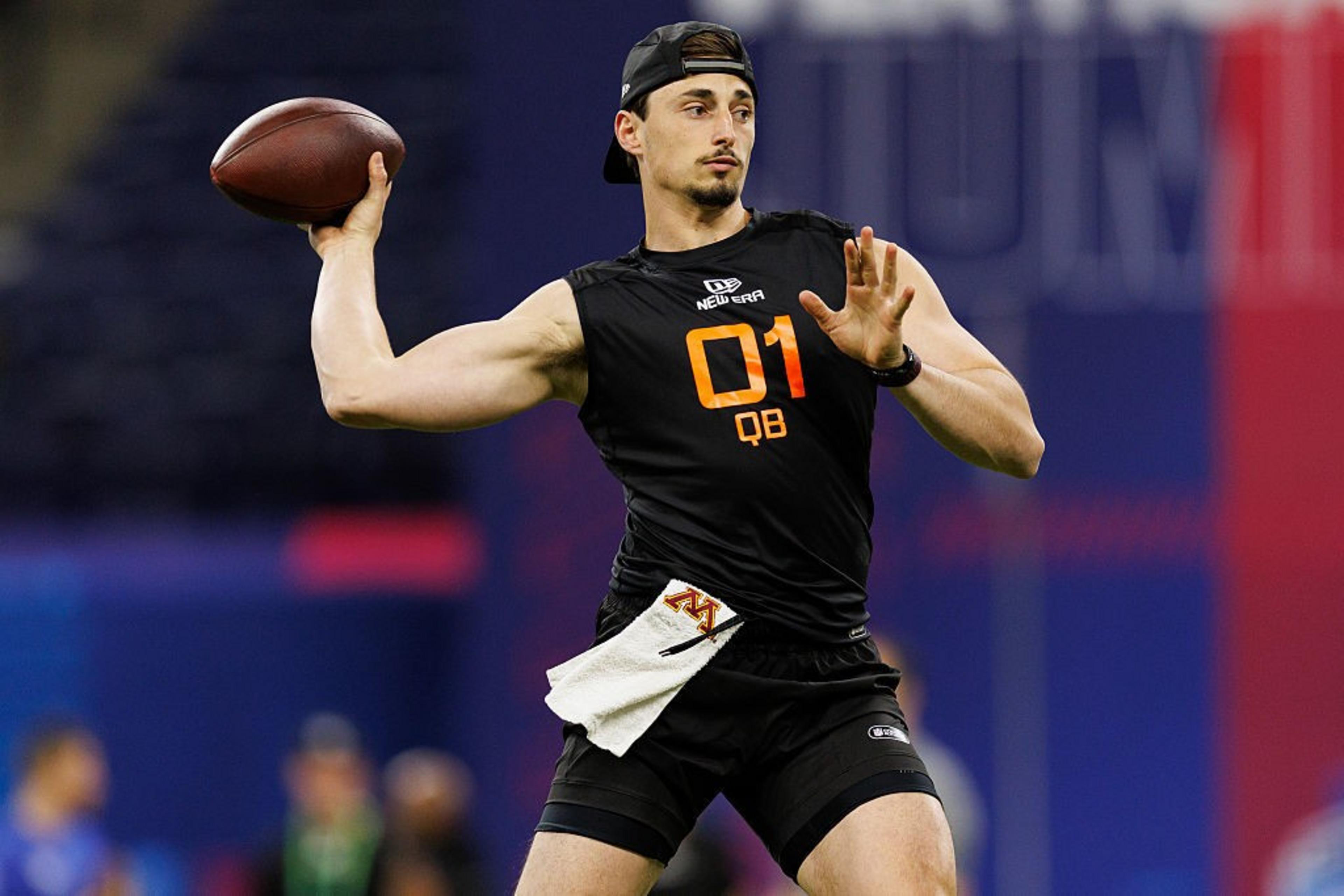 A person in a black athletic outfit with &quot;01 QB&quot; written on it is poised to throw a football. They wear a backward cap and have a towel tucked into their waistband.