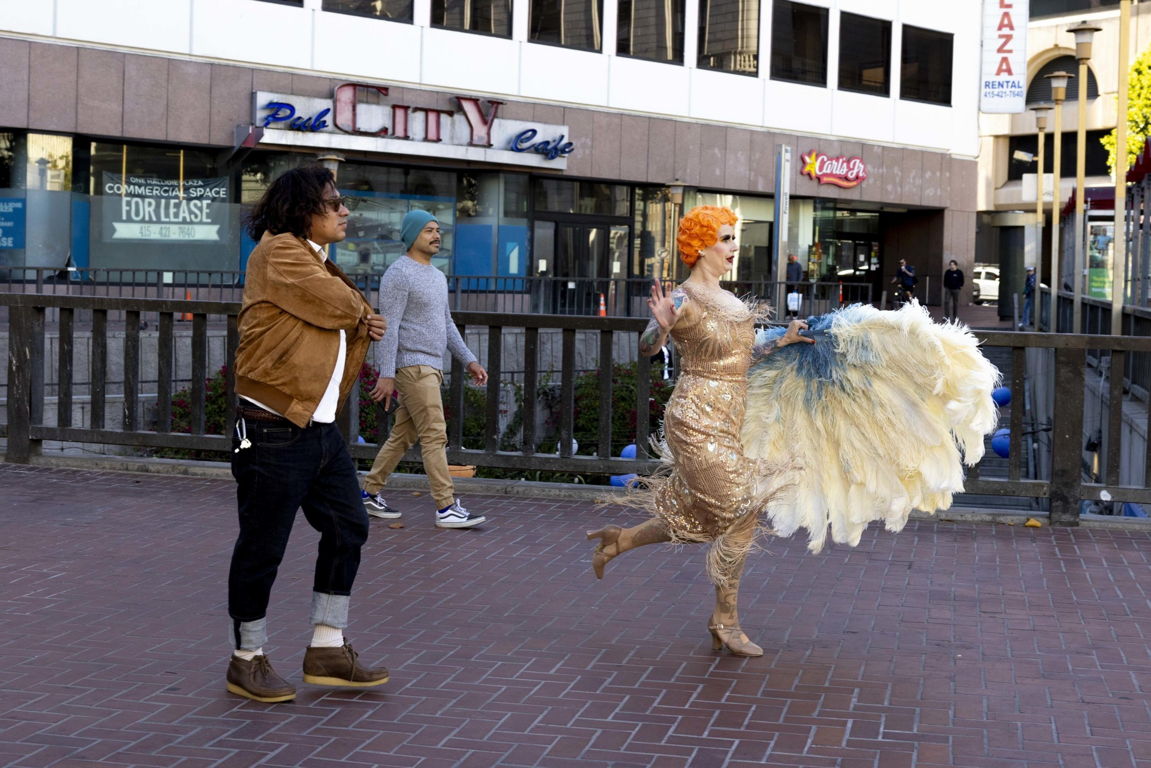 Three people are walking outdoors: one man in a brown jacket, another in a blue hat, and a woman in a sparkling dress carrying large, feathered fans.