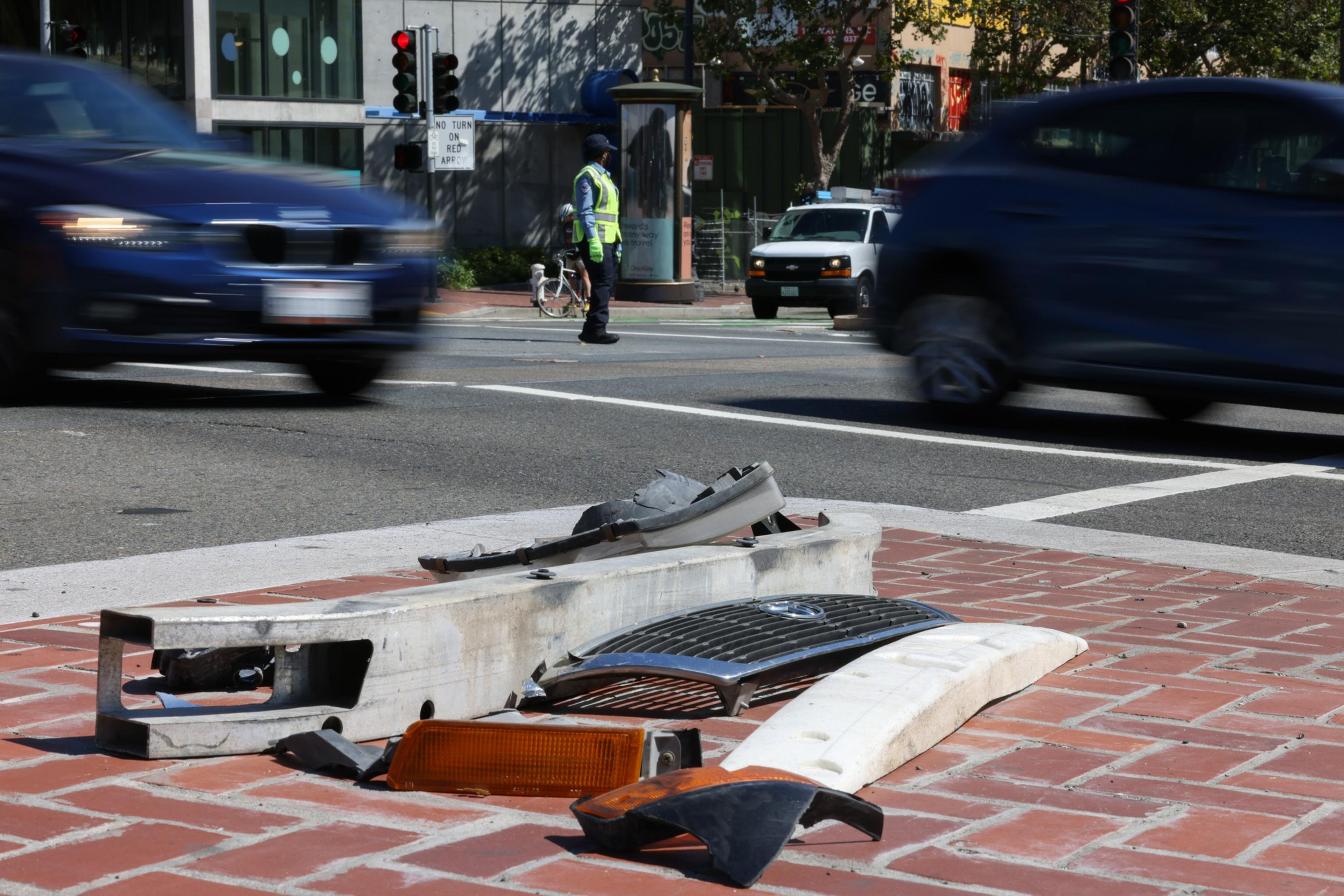 Broken car parts lie on a brick surface at an intersection while cars drive by and a police officer directs traffic in the background.