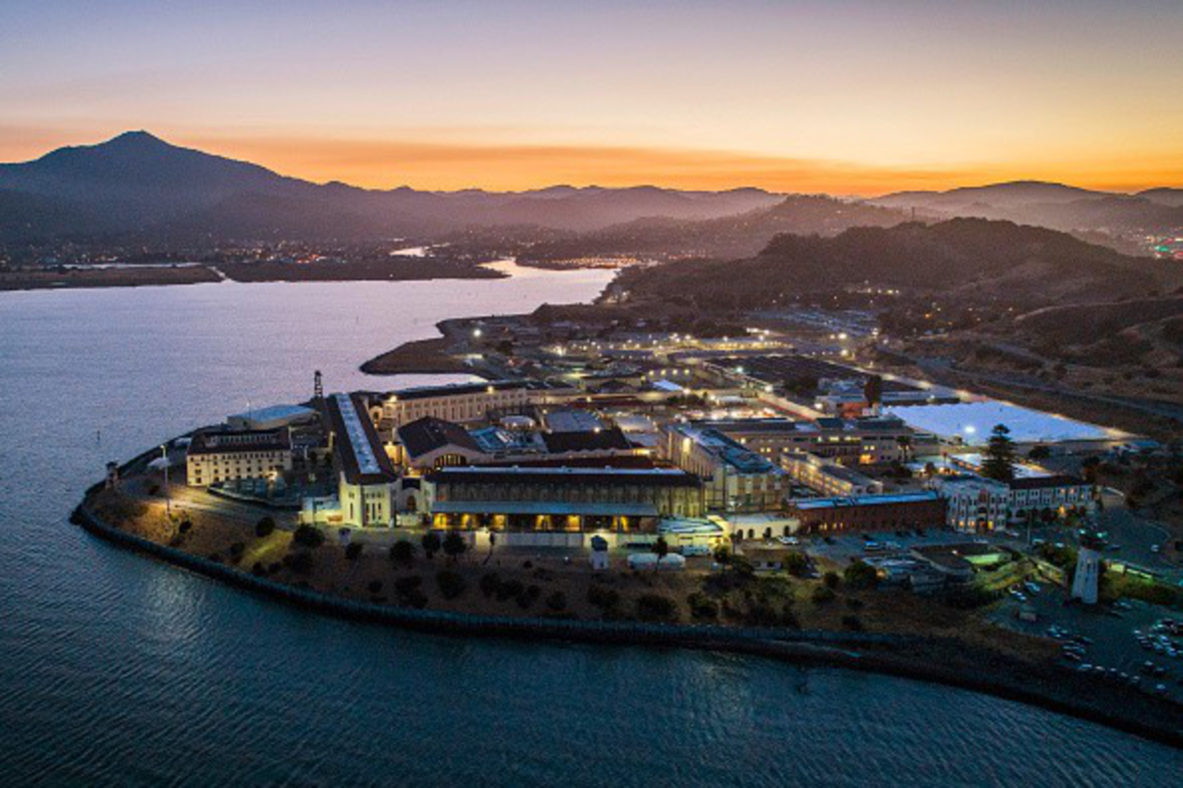 A large island complex with buildings lit up at dusk, surrounded by water, with hills and a colorful sunset sky in the background.
