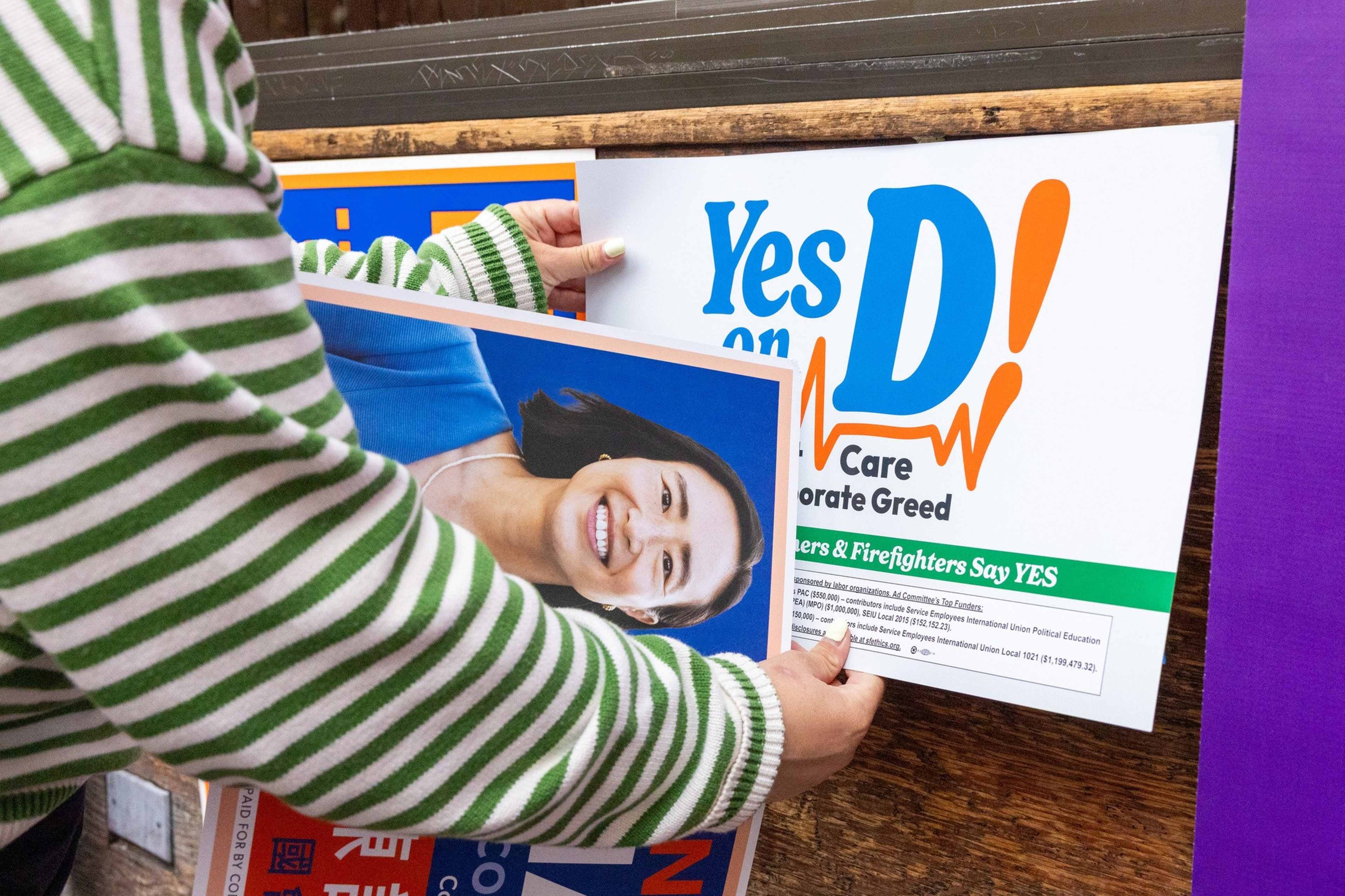 A person in a green and white striped sweater is holding a smiling woman’s poster and placing a “Yes on D!” campaign sign advocating for workers and firefighters.
