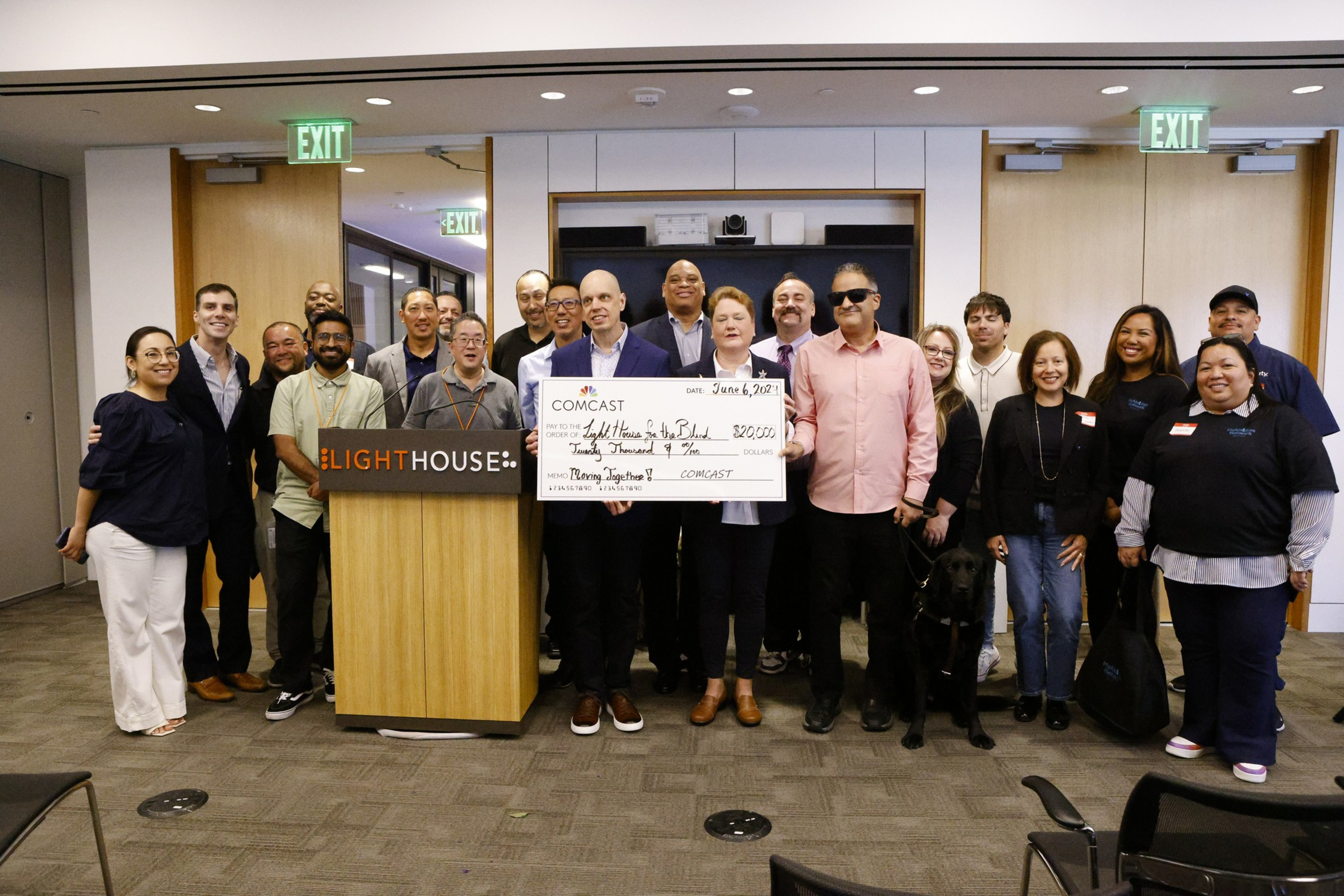 A crowd of people stand at a podium as a giant check from Comcast is awarded to Lighthouse for the Blind.