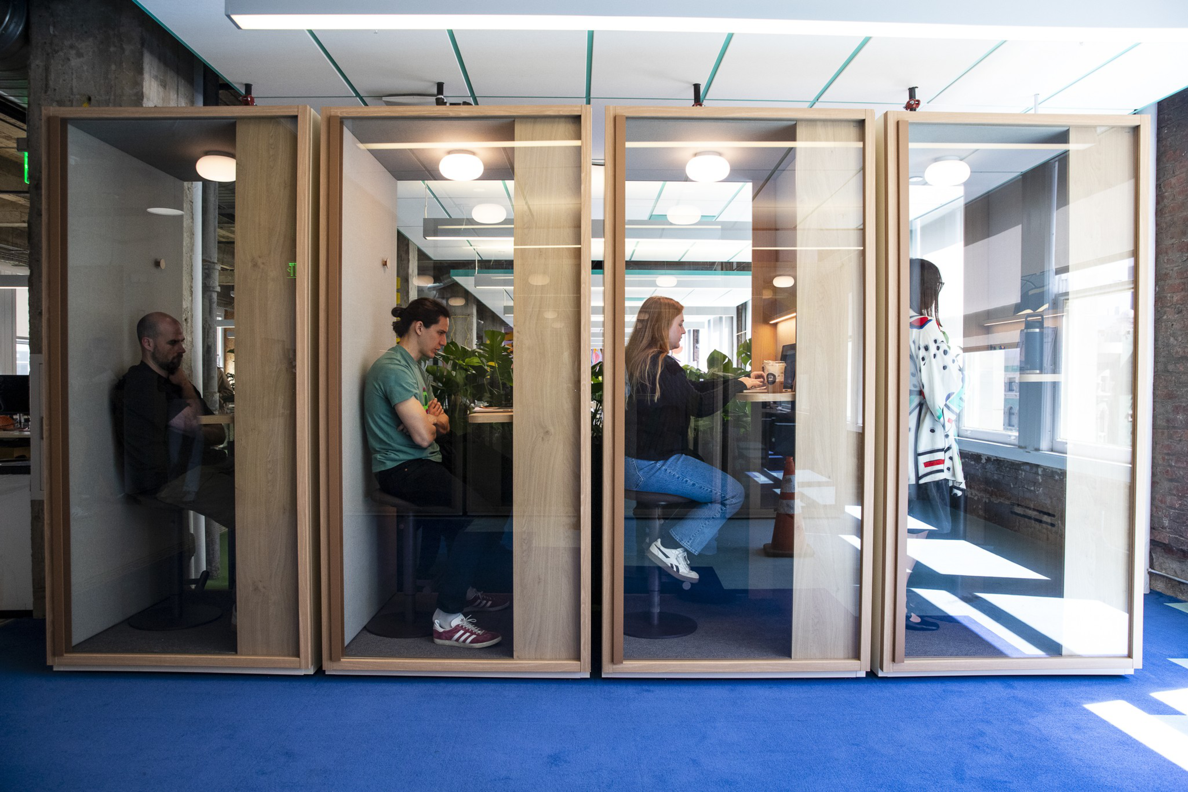 Four people sit or stand separately in individual glass phone booths in a modern office space with blue carpet and bright overhead lighting.