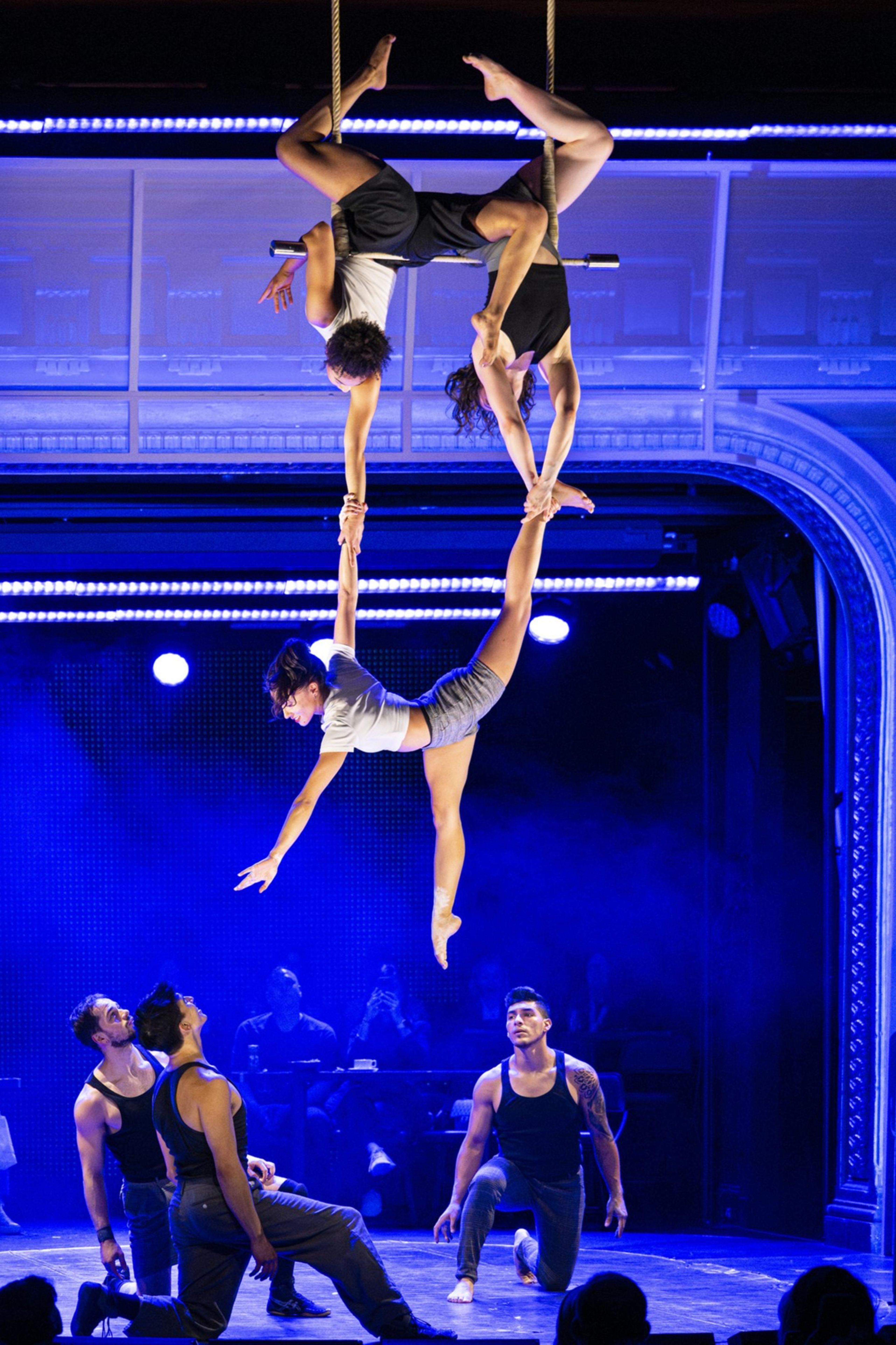 Acrobats perform on stage; two hang upside down, holding a third person with their hands. Below, three men kneel, watching. Blue lighting highlights the scene.