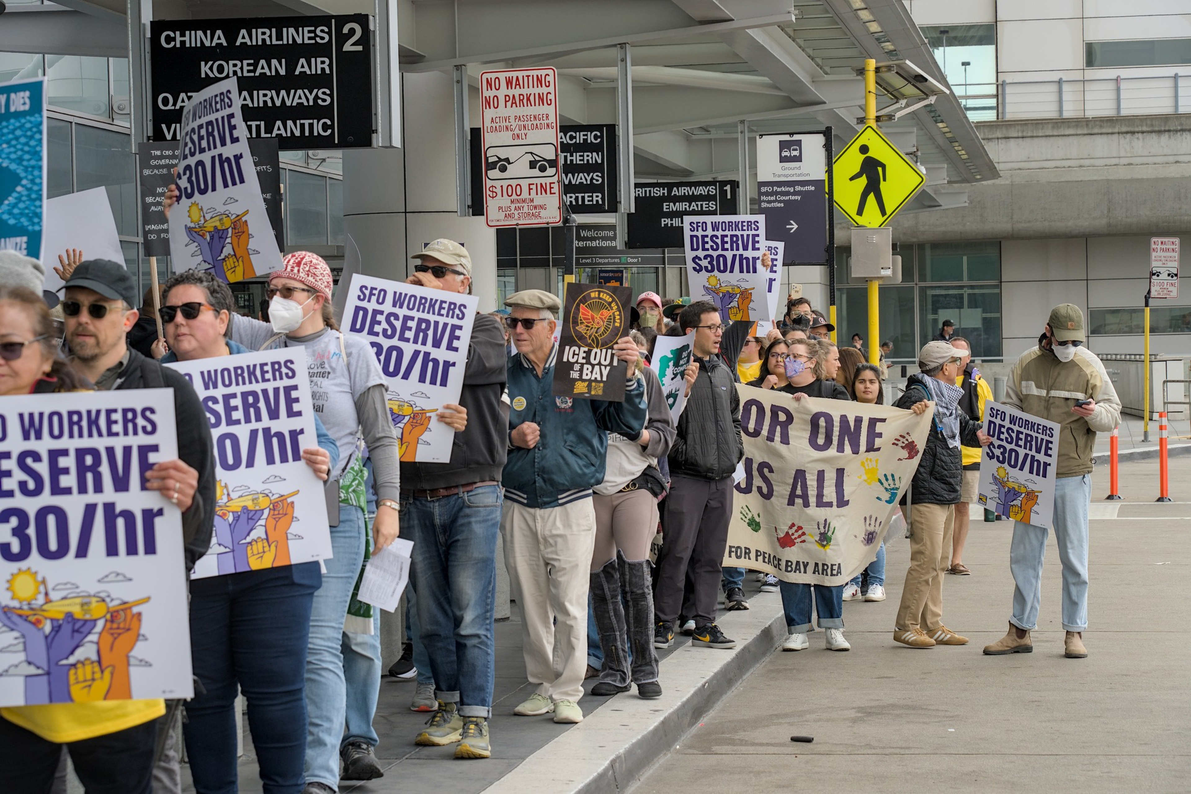 People line up outdoors holding signs that say “SFO WORKERS DESERVE $30/hr” and “FOR ONE IS ALL,” protesting for better wages at an airport terminal.