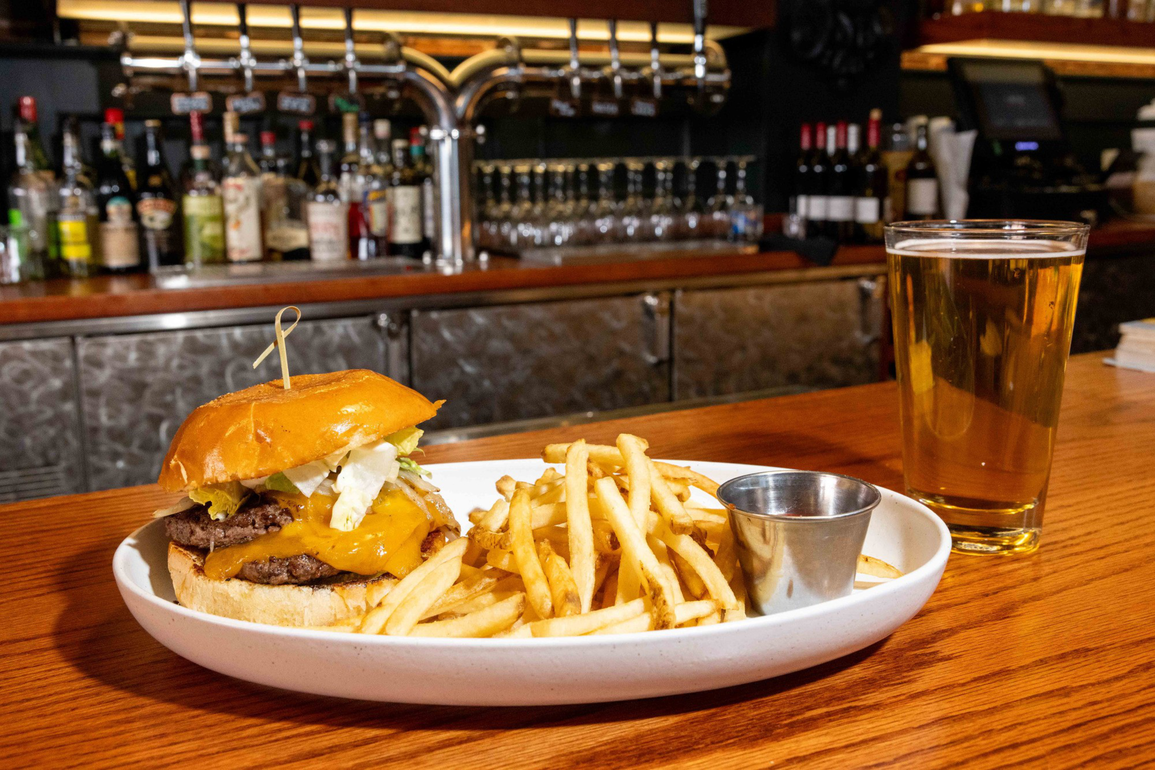 A cheeseburger with lettuce and a side of fries sit on a wooden bar counter, alongside a pint of beer, with liquor bottles visible in the background.