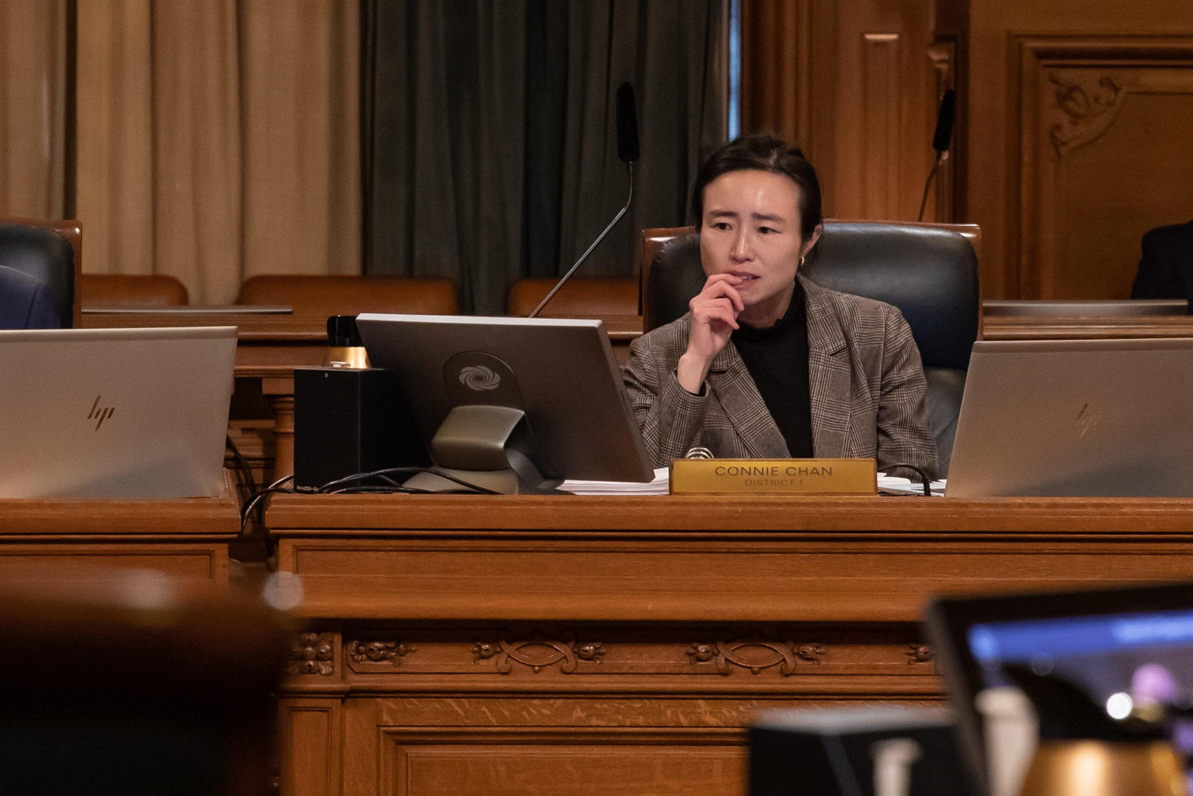 A woman in a brown plaid blazer sits at a wooden desk with two laptops and a nameplate that reads "Connie Chan." She appears to be thinking.