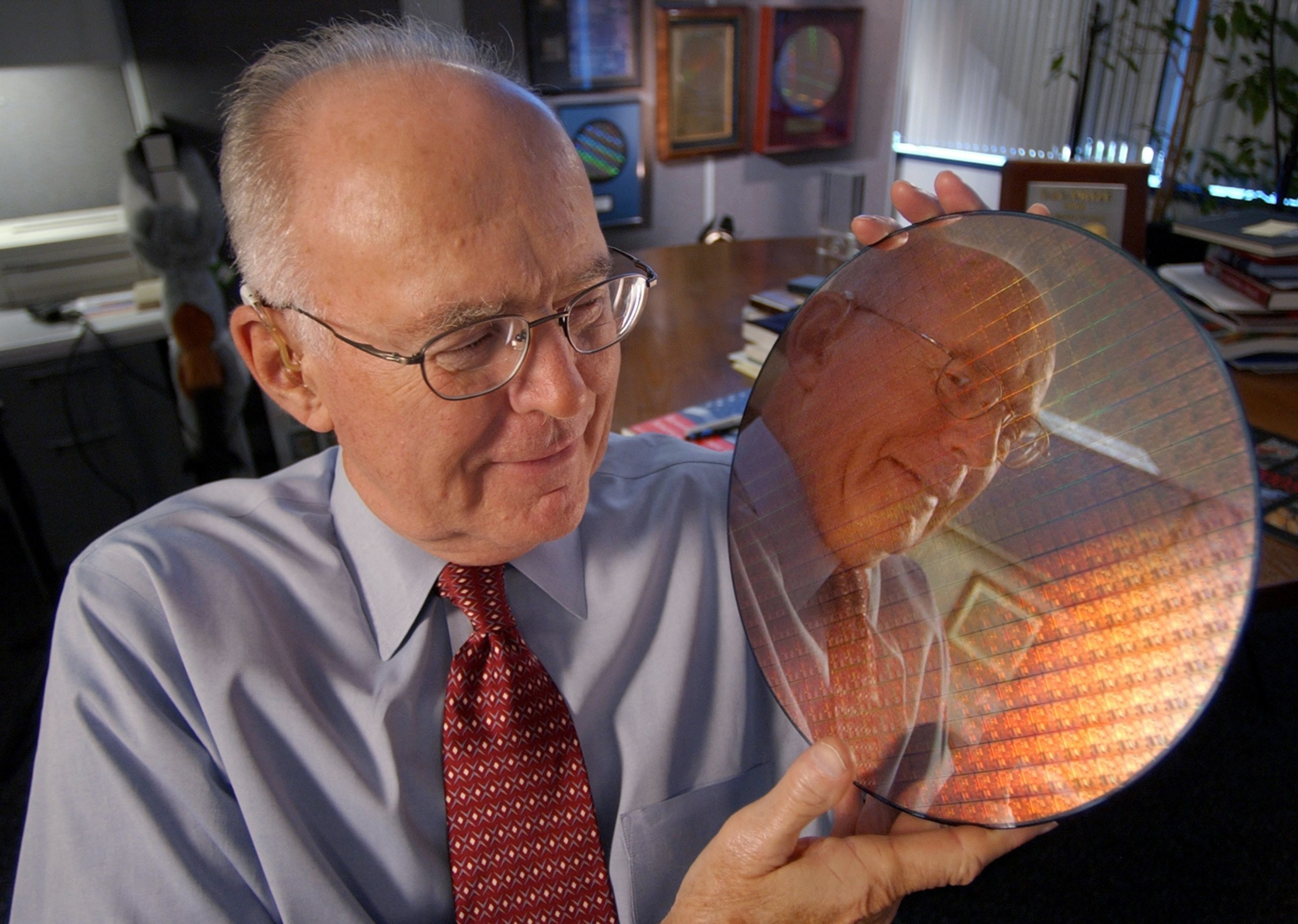An elderly man wearing glasses and a tie holds a reflective, circular object, showing his reflection. The background features framed items and a well-lit room.