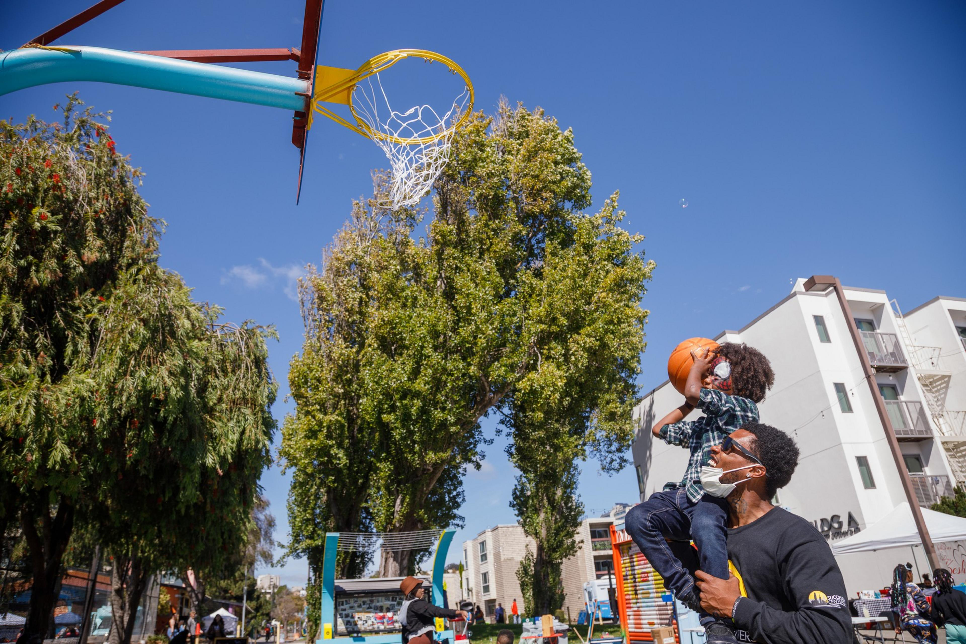 An adult lifts a child towards a basketball hoop, as the child aims a ball for the shot, outdoors under a clear blue sky.