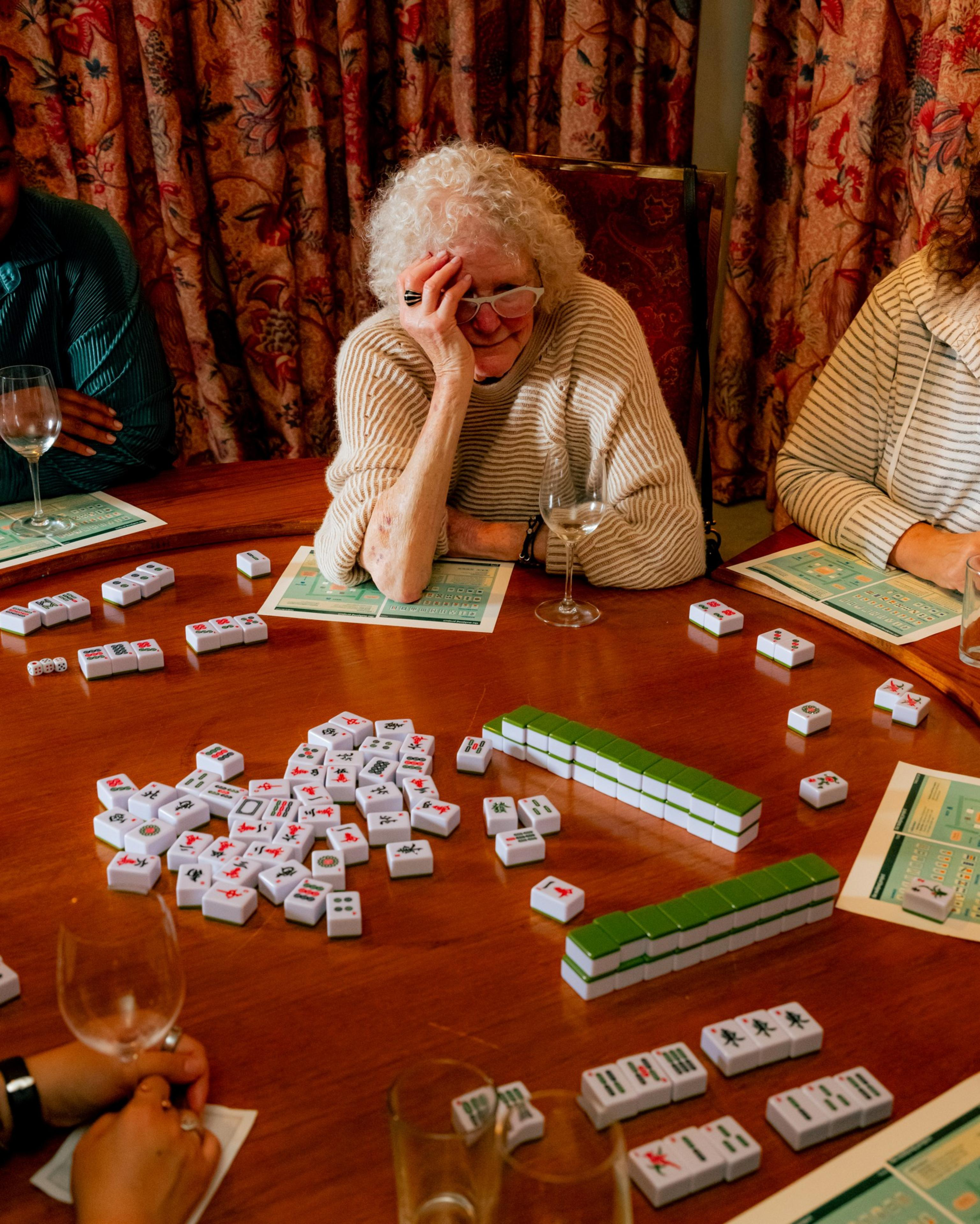An older woman with curly white hair and a striped sweater leans on a table playing Mahjong with others, with tiles, score sheets, and wine glasses scattered around.