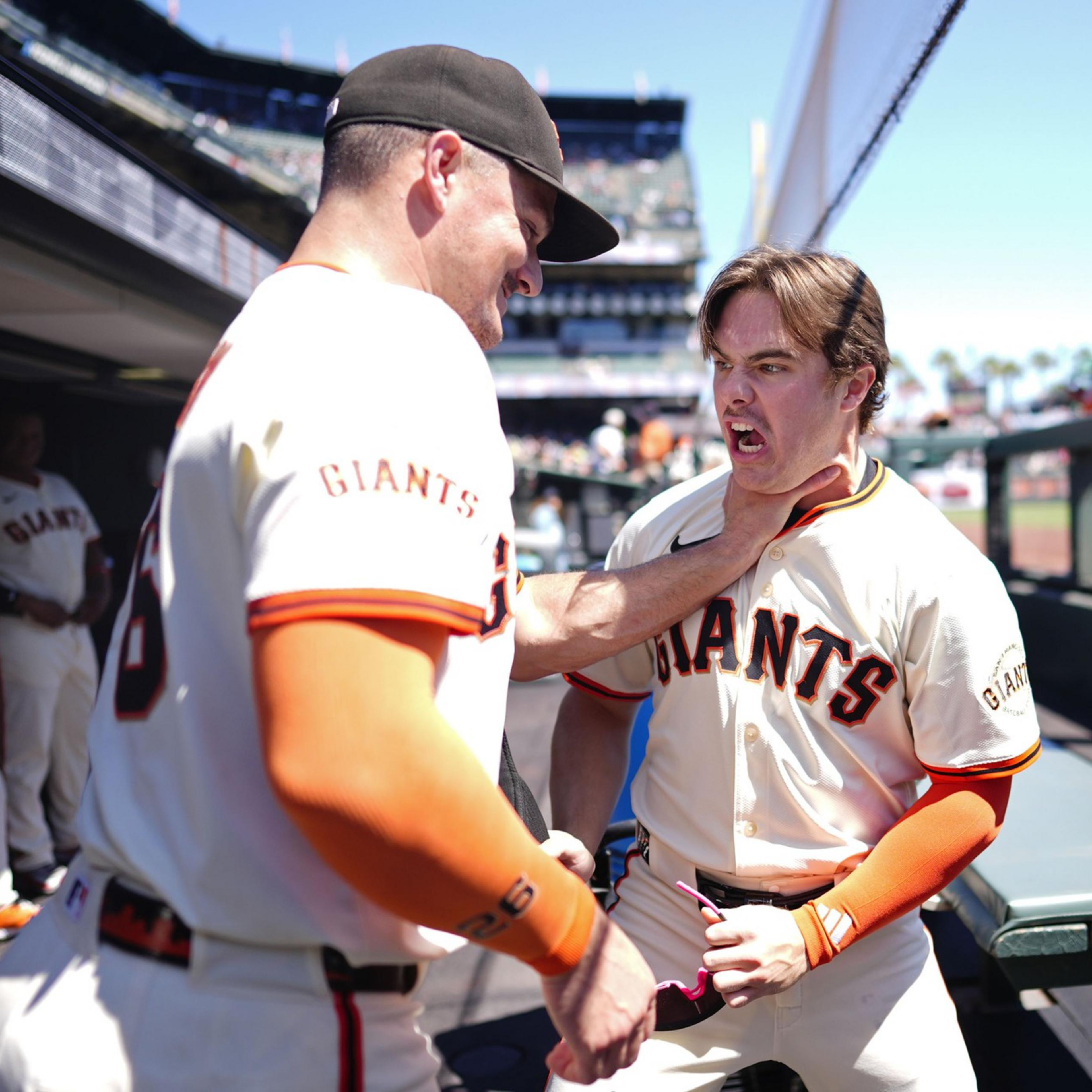 A baseball player in a Giants uniform pretends to choke a teammate while another player watches nearby in the dugout under a clear sky.