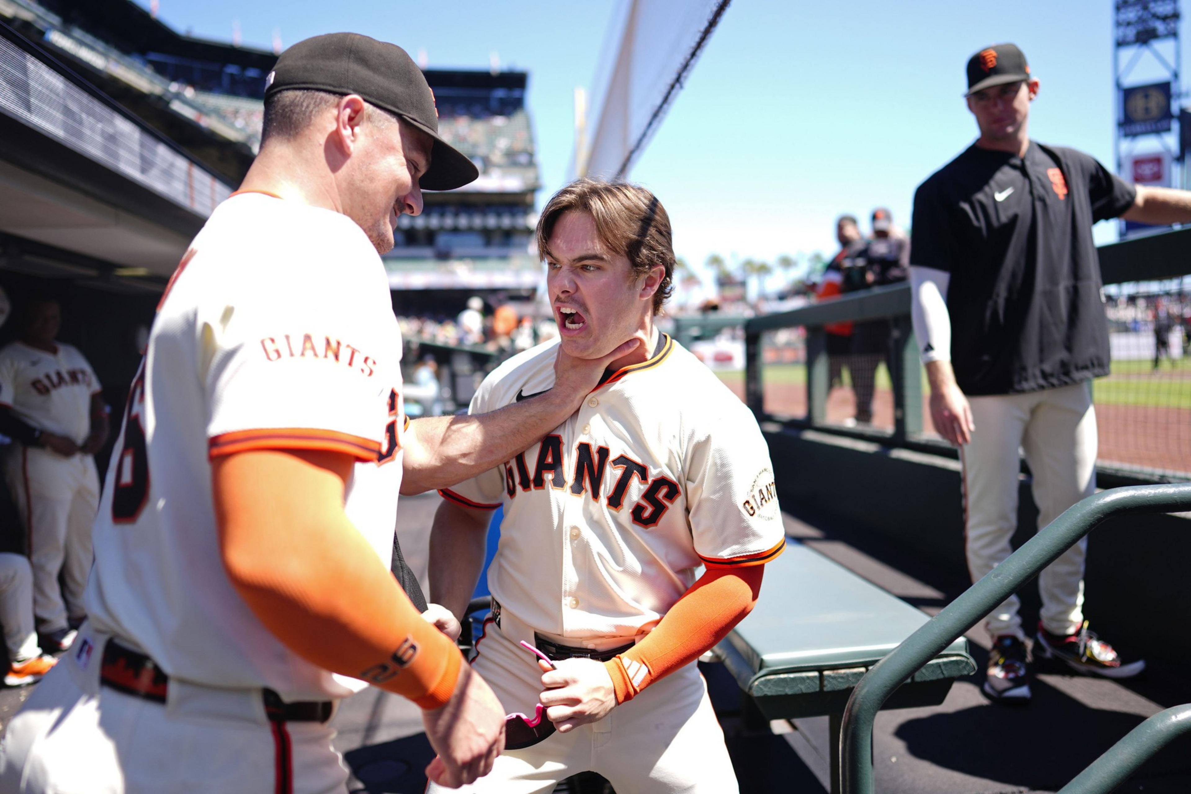 A baseball player in a Giants uniform pretends to choke a teammate while another player watches nearby in the dugout under a clear sky.