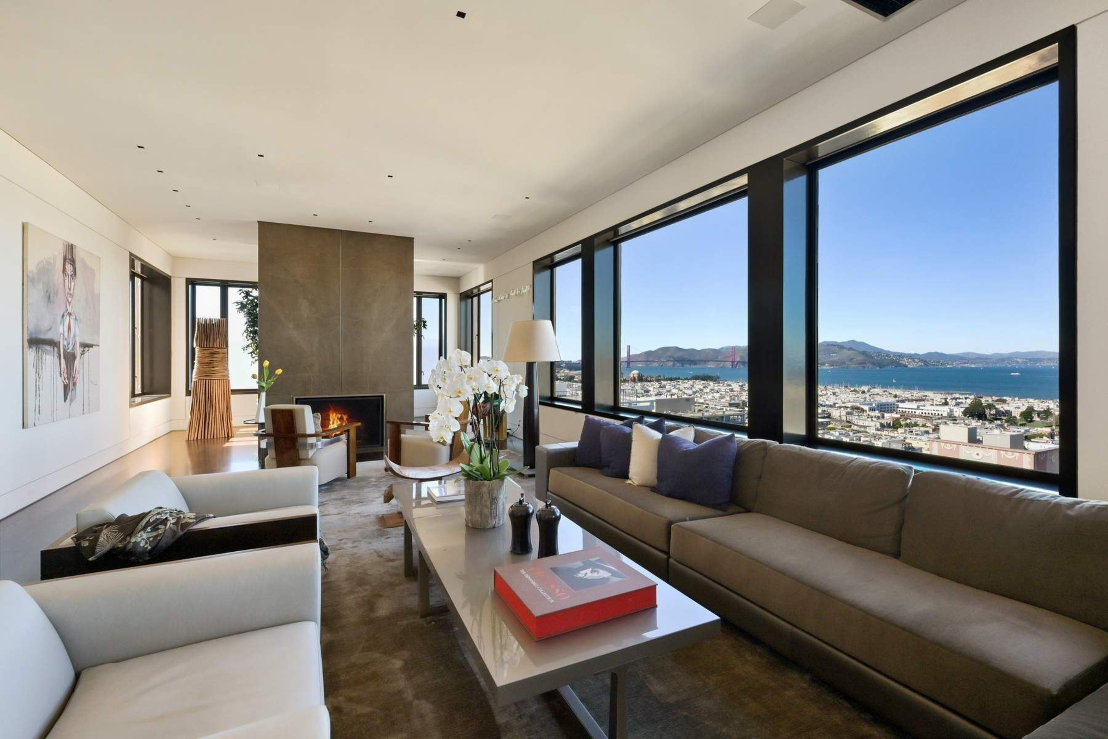 A modern living room with large windows overlooking a city and bay, featuring a long sofa, armchairs, a coffee table, fireplace, and decorative flowers.