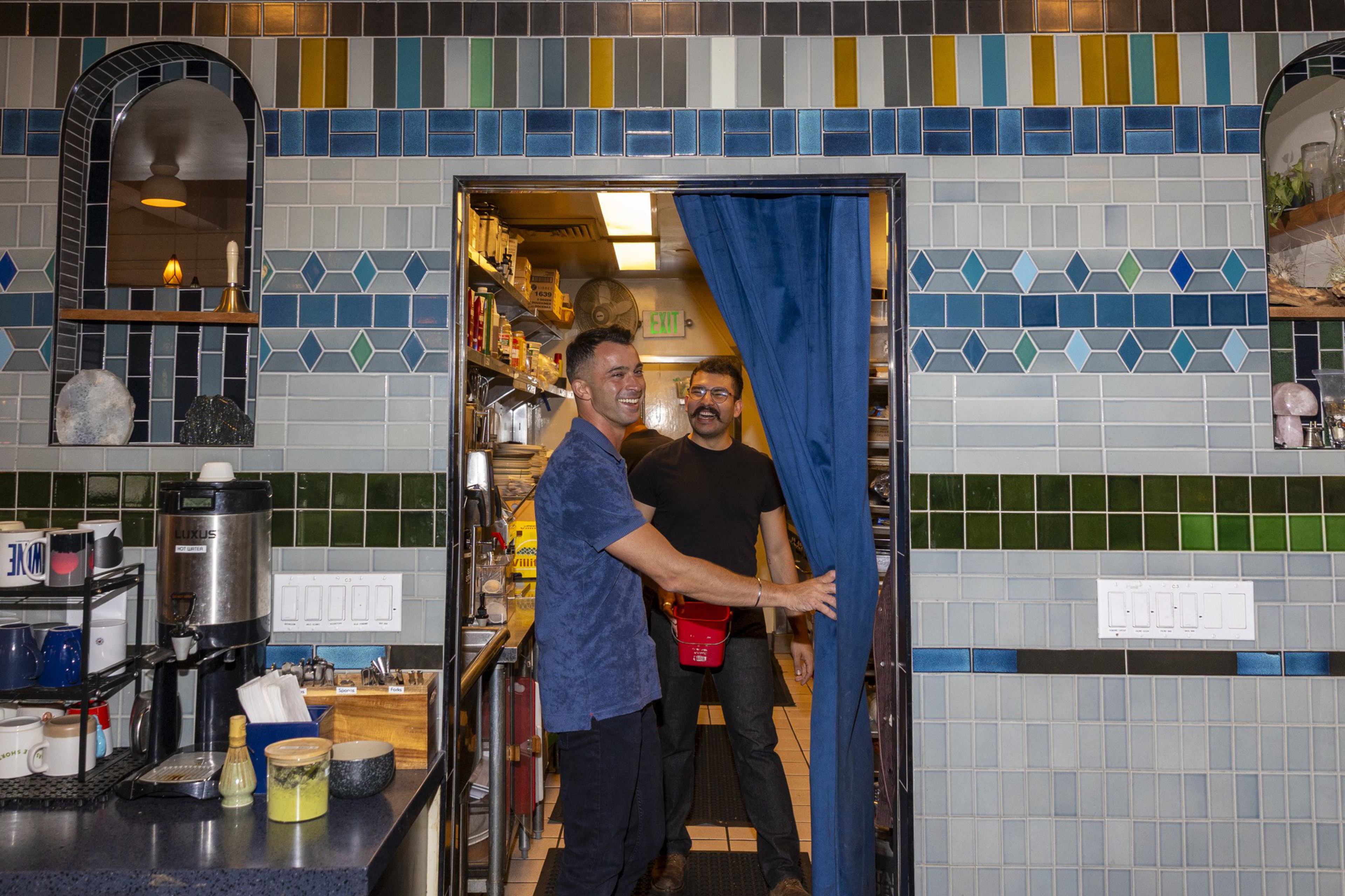 Two men smile while standing in a doorway with a blue curtain, inside a kitchen area with patterned tile walls and a coffee station nearby.
