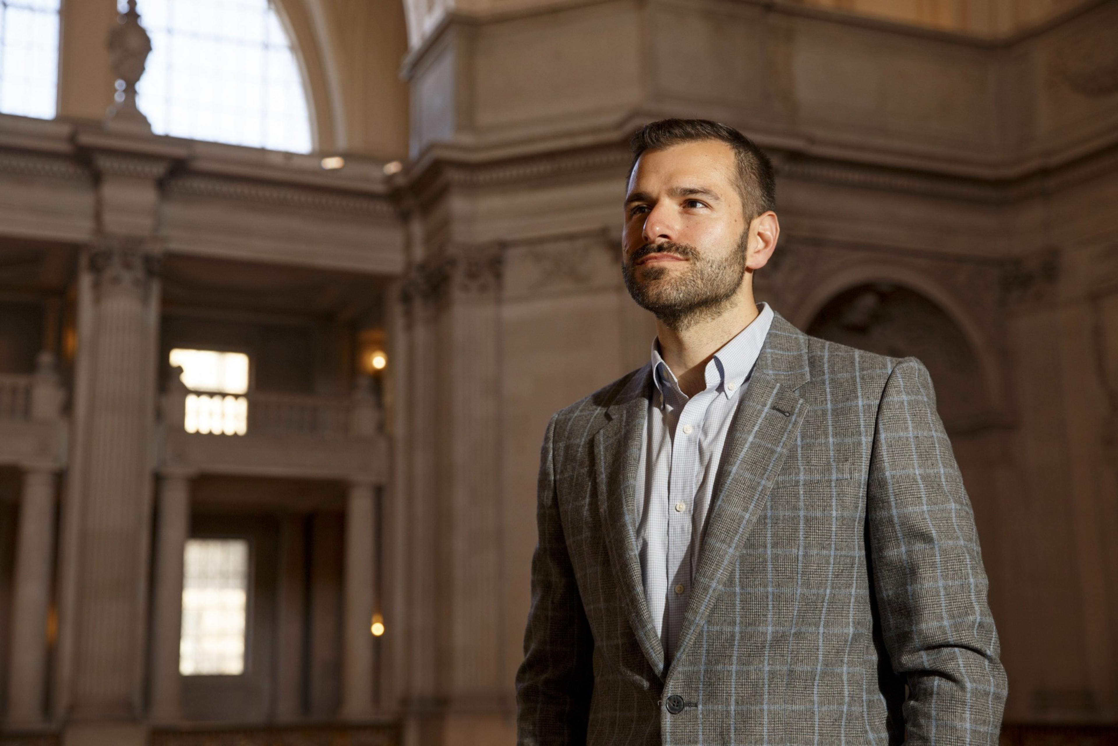 A man in a checkered blazer stands confidently in an elegant, sunlit hall with classical architecture.