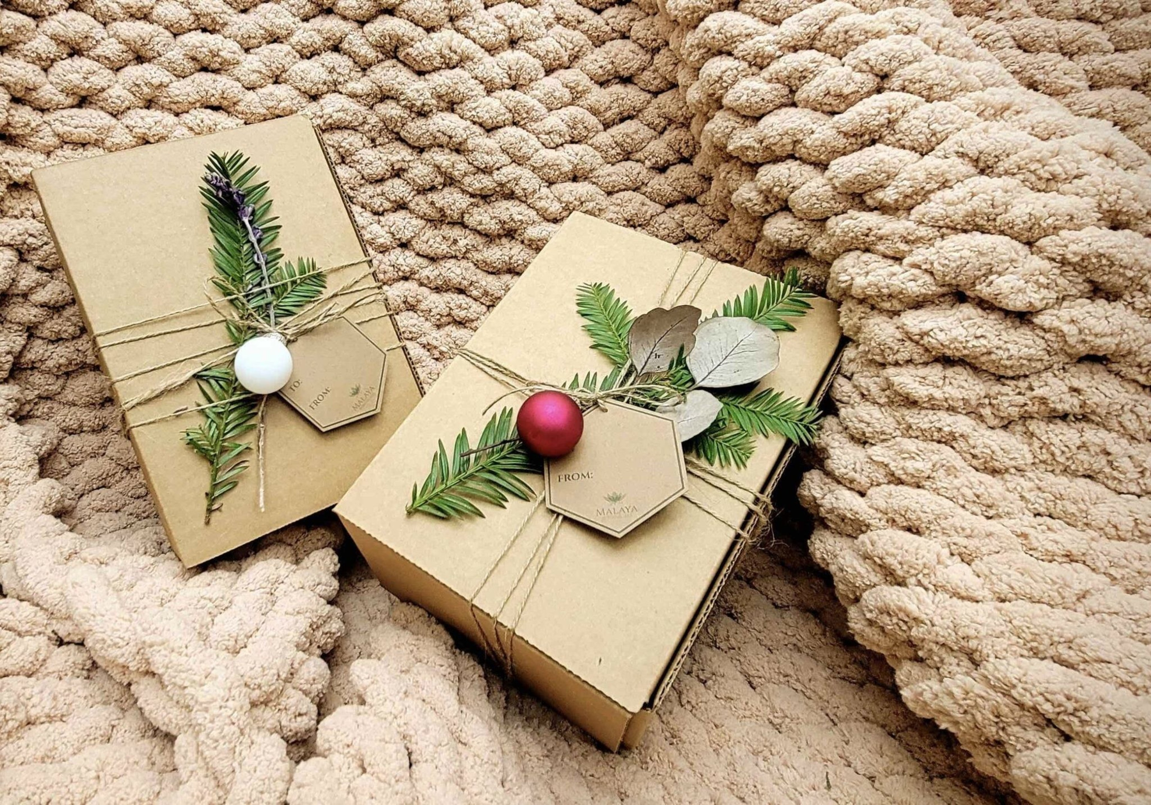 Two gift boxes wrapped in brown paper and tied with twine are decorated with evergreen sprigs, small leaves, and red and white baubles, resting on a chunky knit blanket.