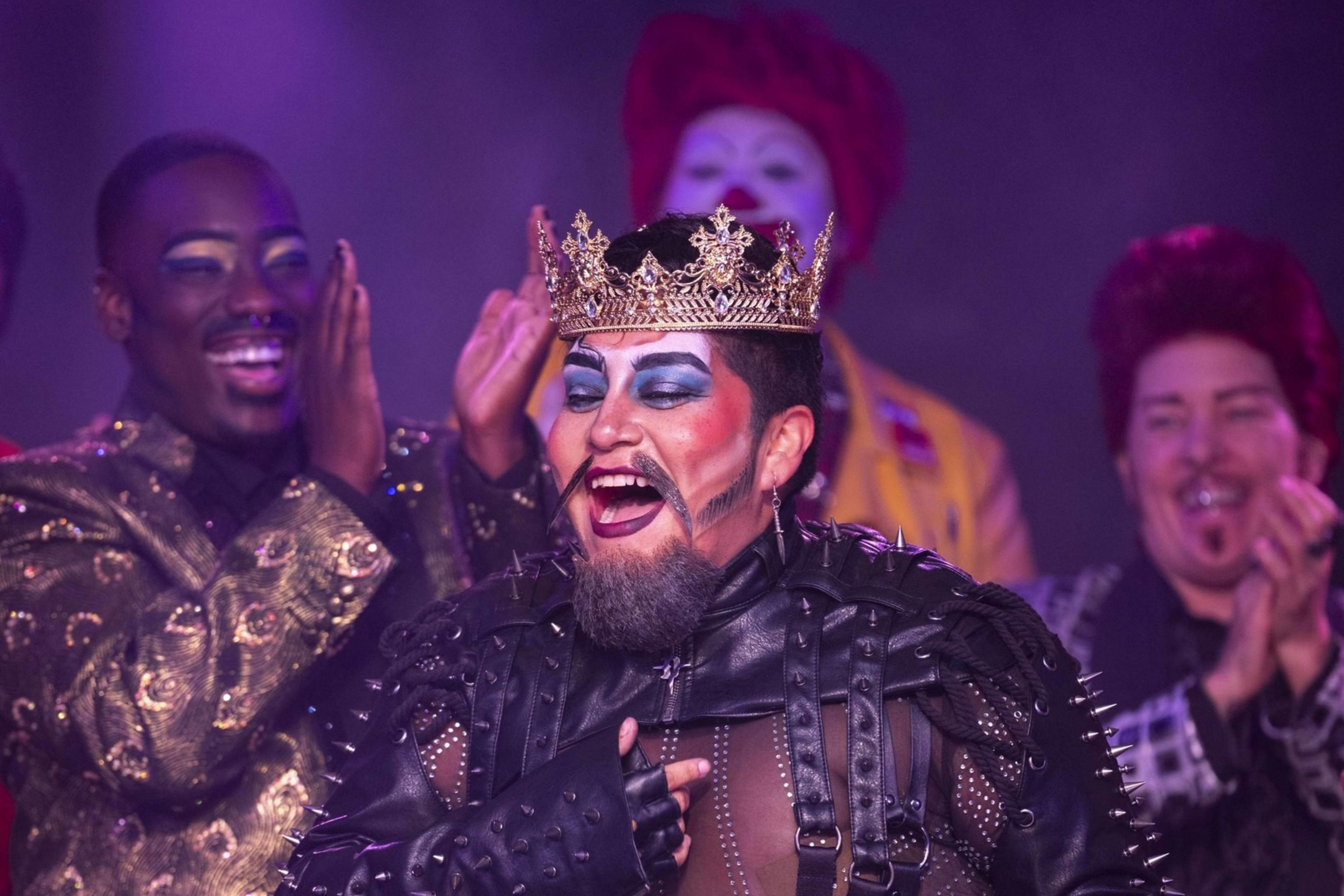 A person in a studded leather outfit and crown jubilantly holds a hat while being applauded by a diverse group of extravagantly dressed and makeup-clad individuals.