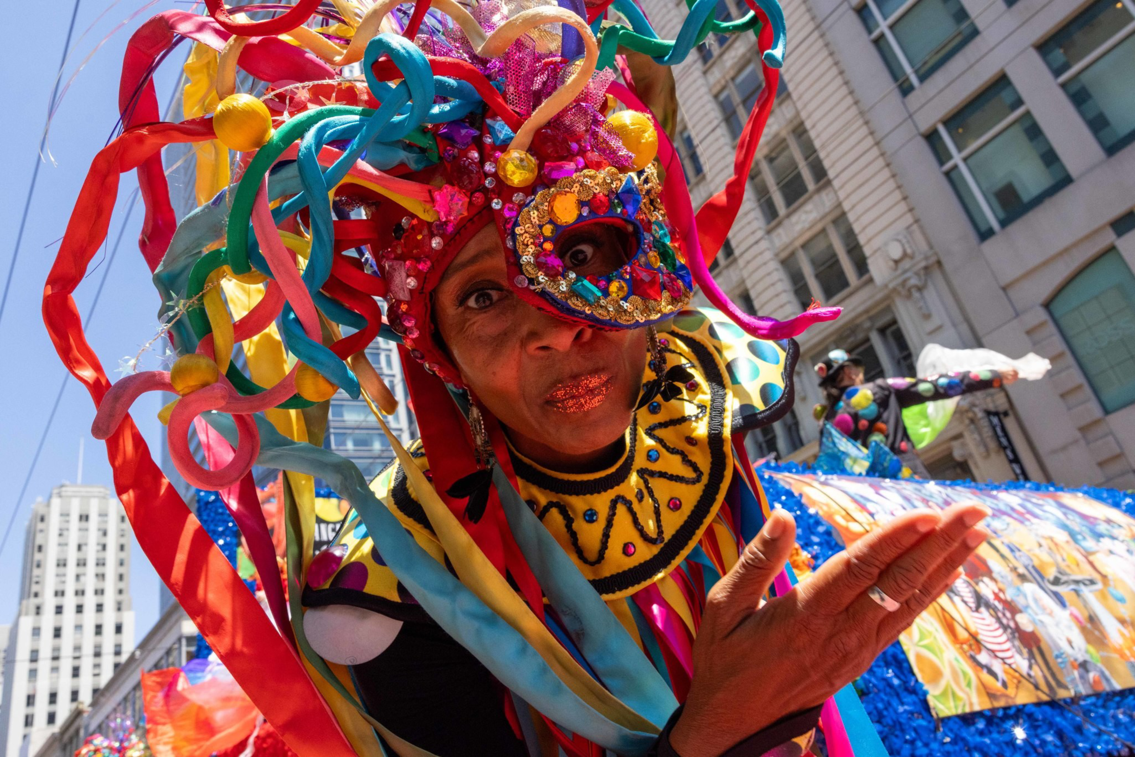 A person in a colorful, elaborate costume with ribbons and a decorated mask is performing in a vibrant street parade in a cityscape setting.