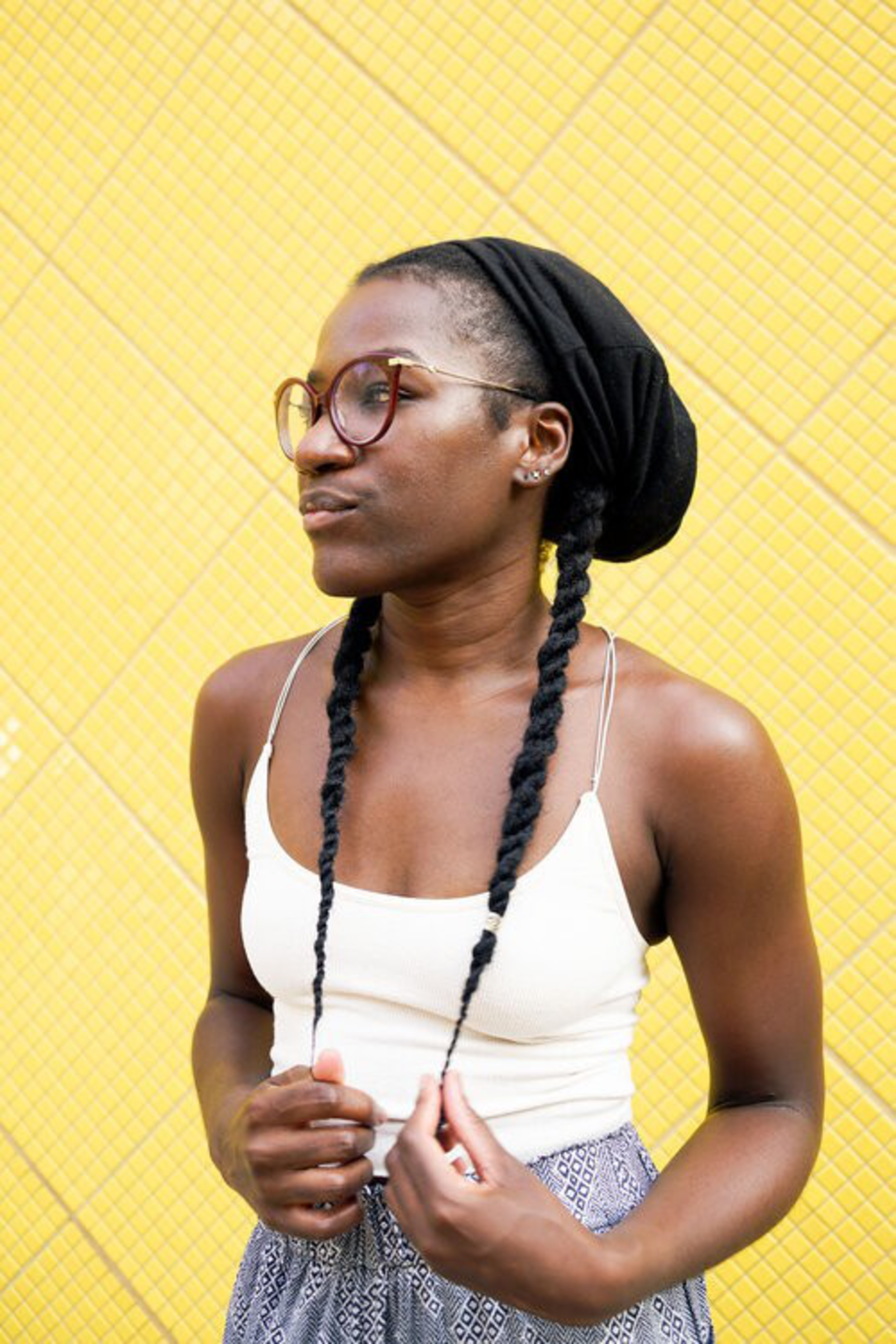 A woman with braided hair and glasses wears a white tank top and patterned skirt, standing against a bright yellow textured wall.